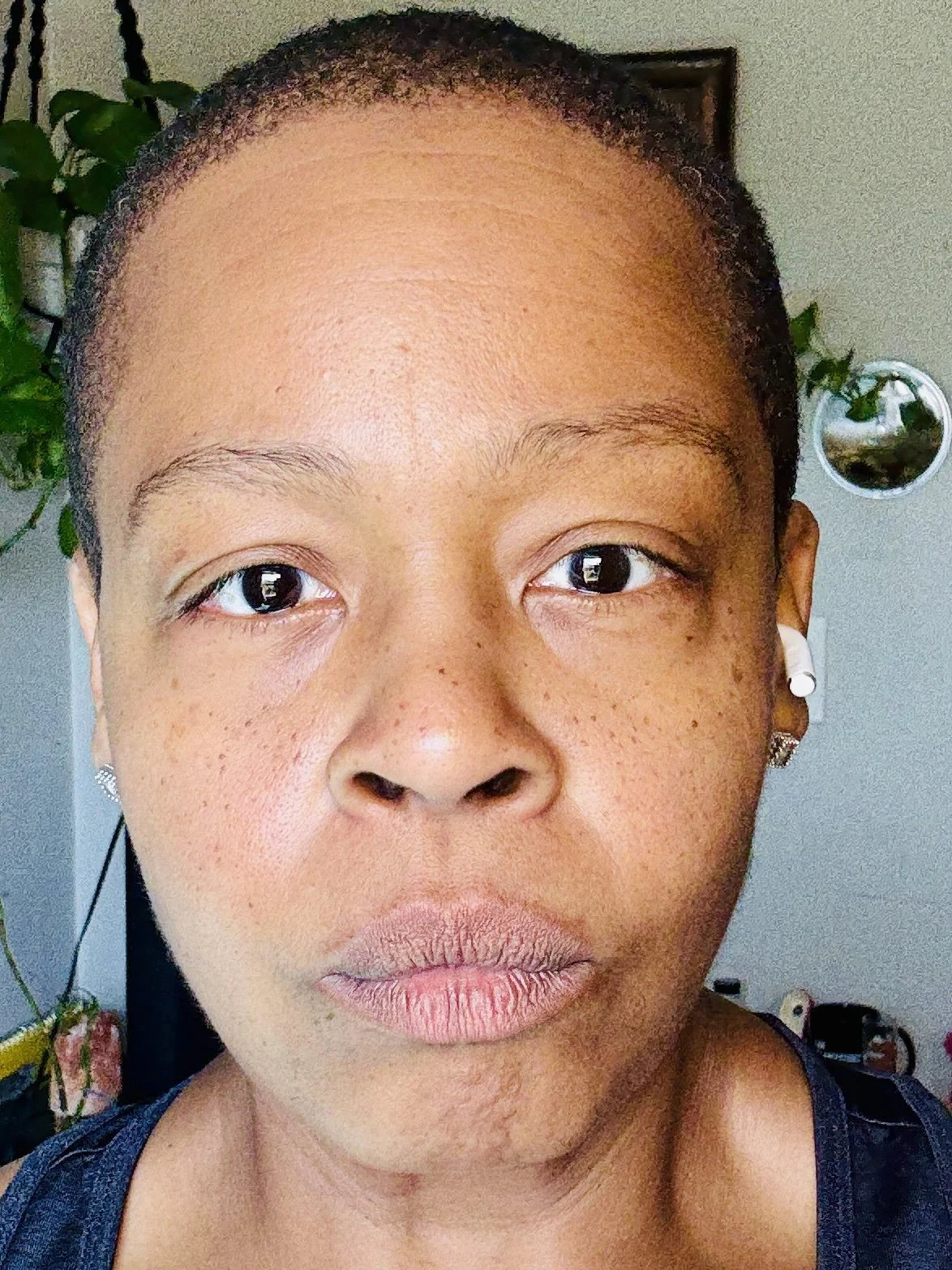 Erica DeLaine Bradshaw with short hair, slightly raised eyebrows, and wearing earrings, with indoor plants and framed picture in the background.