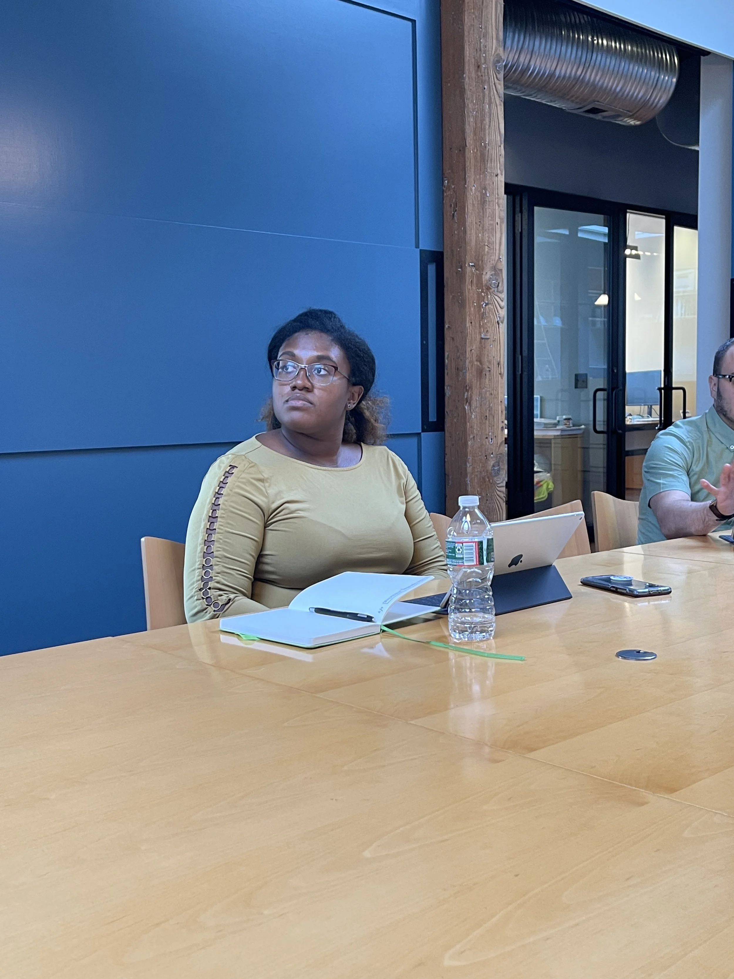 A woman sitting at a conference table with a notebook, pen, water bottle, and tablet in front of her, looking attentively.