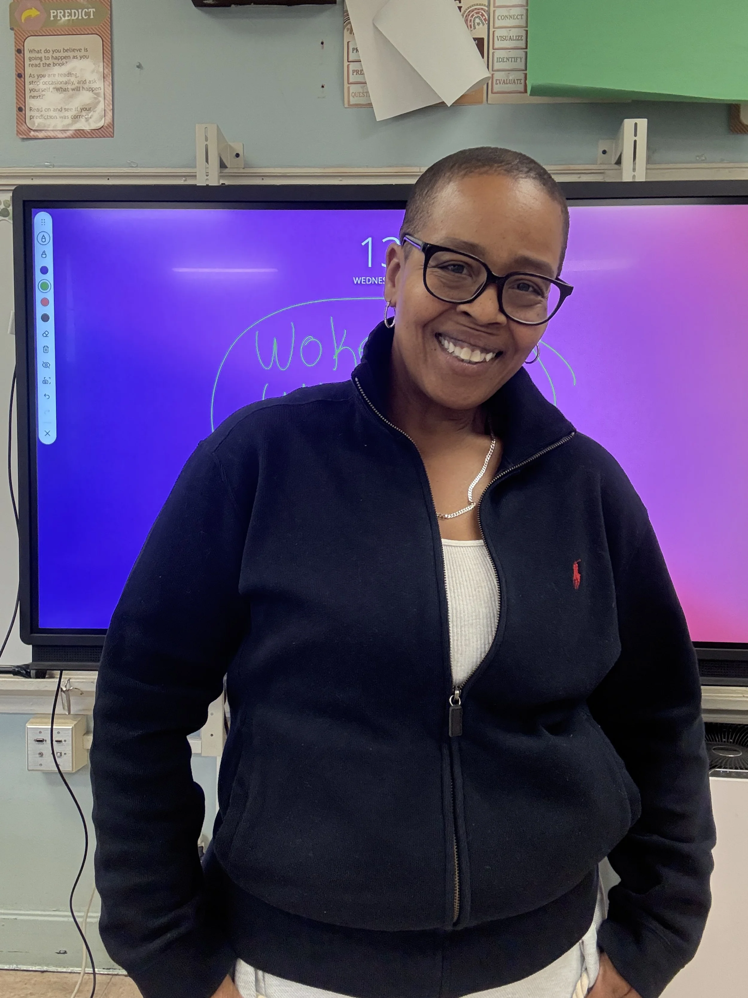 A woman with short hair, glasses, and hoop earrings smiling in front of a digital screen in a classroom. She is wearing a black zip-up jacket over a white shirt.