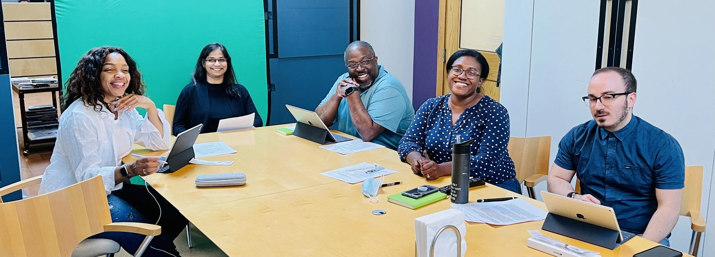 Group of five diverse people smiling and sitting around a conference table with laptops, papers, and drinks in a meeting room.