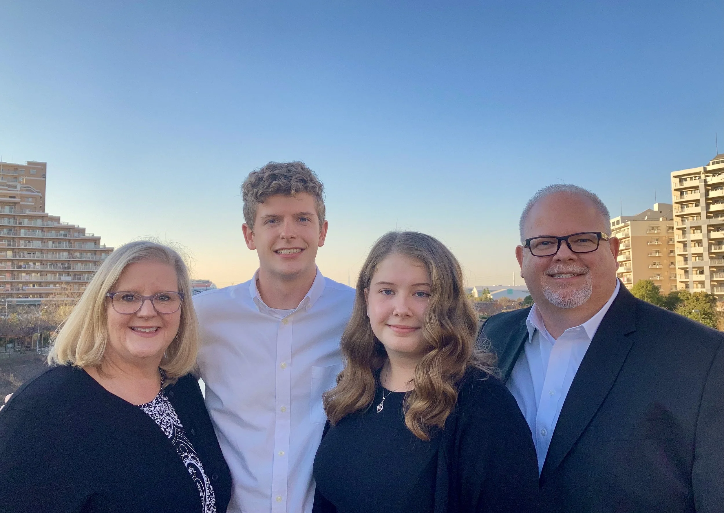 A family of four posing outdoors with city buildings and a blue sky in the background. The family consists of a woman with blonde hair and glasses, a young man with curly hair in a white shirt, a young woman with long wavy hair in a black top, and a man with glasses and a beard in a suit jacket and white shirt.