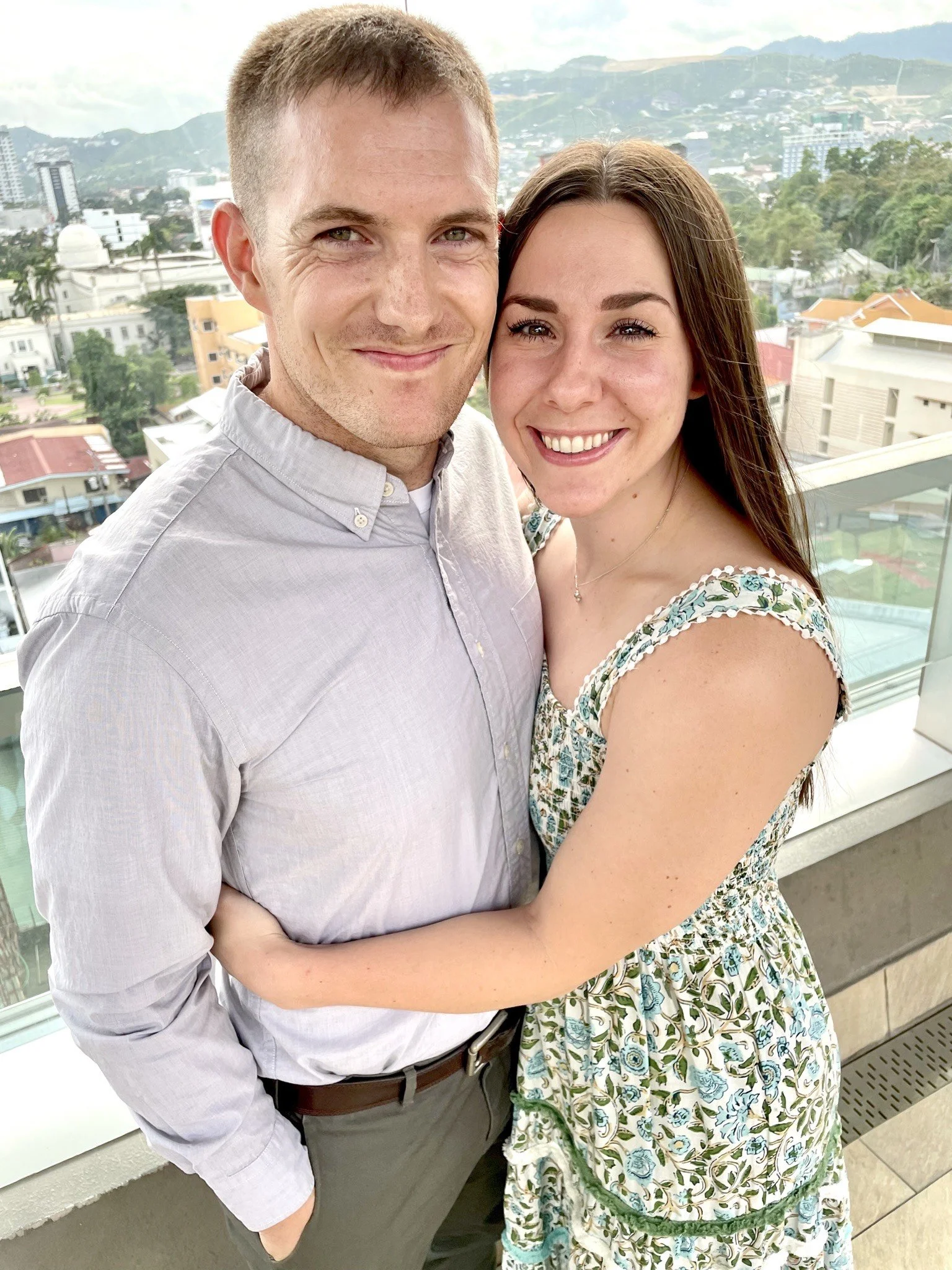 A smiling couple standing on a balcony with a cityscape and mountains in the background.