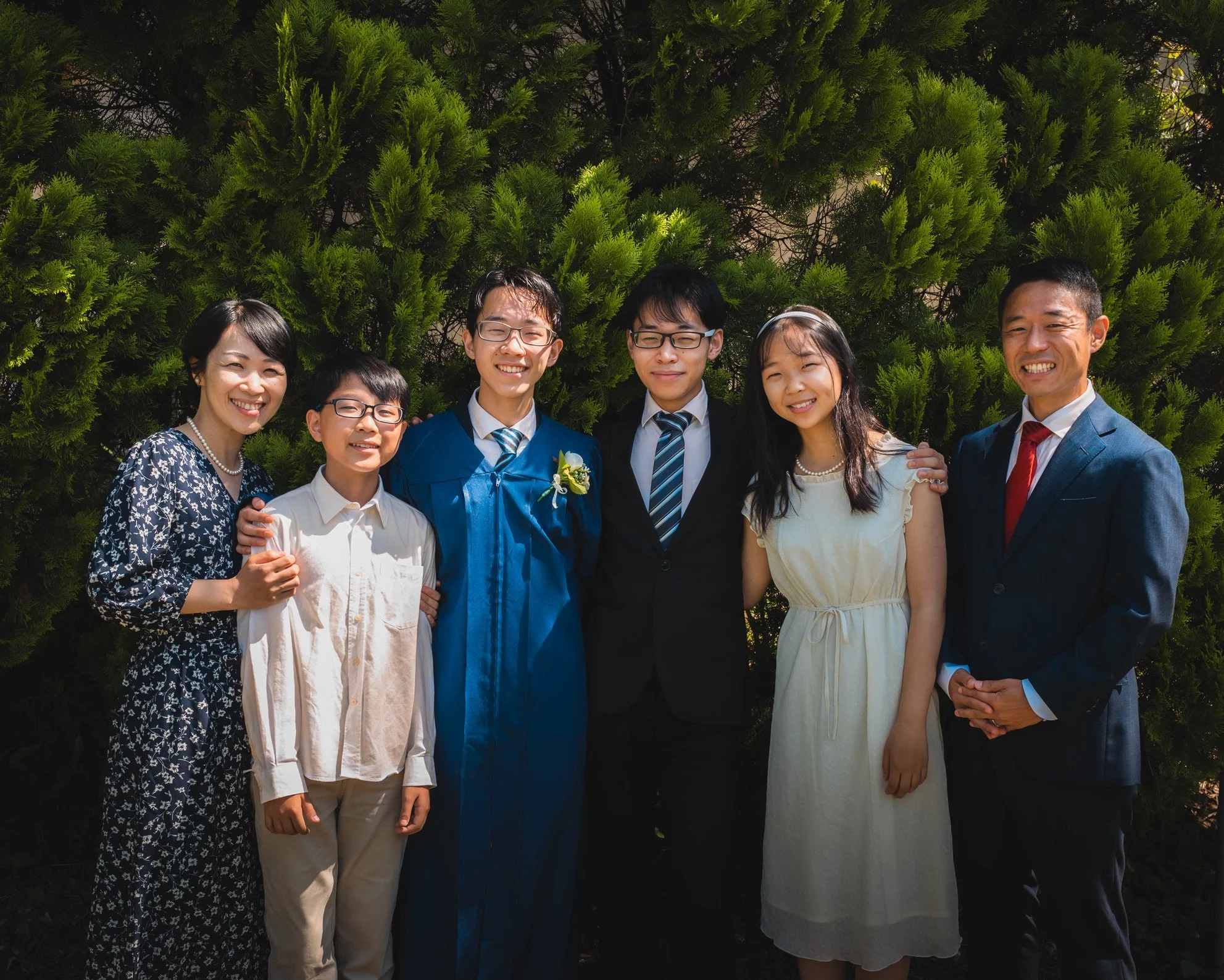 Family celebrating graduation outdoors, standing in front of green bushes, wearing formal and semi-formal attire.