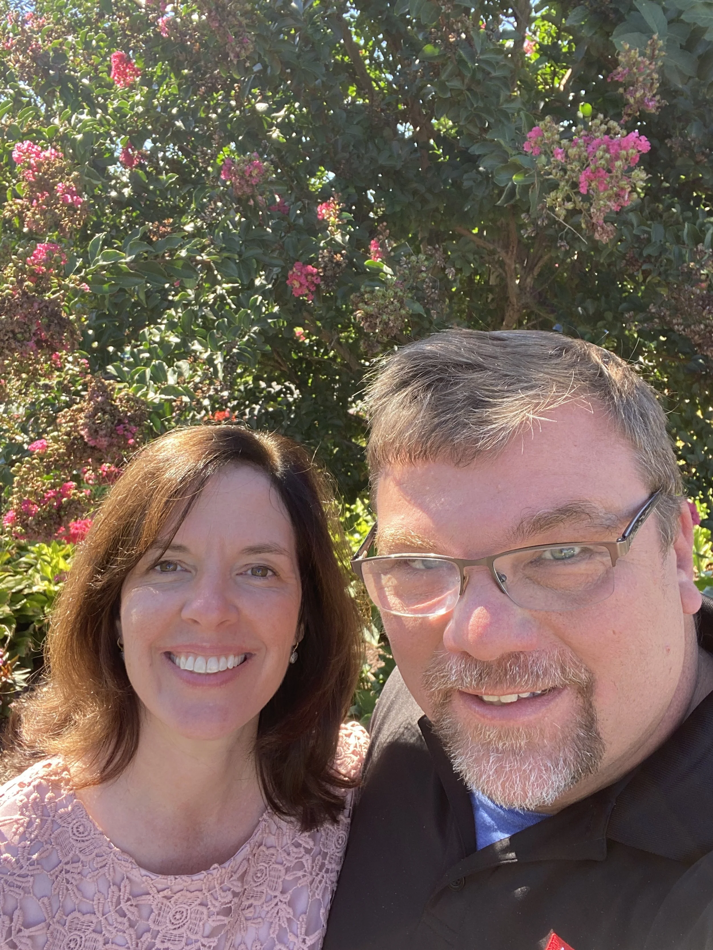 A smiling woman with shoulder-length brown hair and a man with glasses and a beard taking a selfie outdoors in front of a flowering bush with pink and purple flowers.