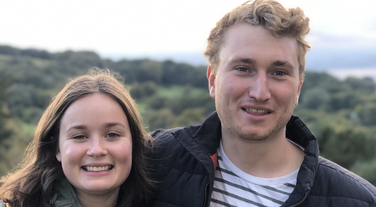 A young woman and a young man smiling outdoors with a blurred green landscape in the background.