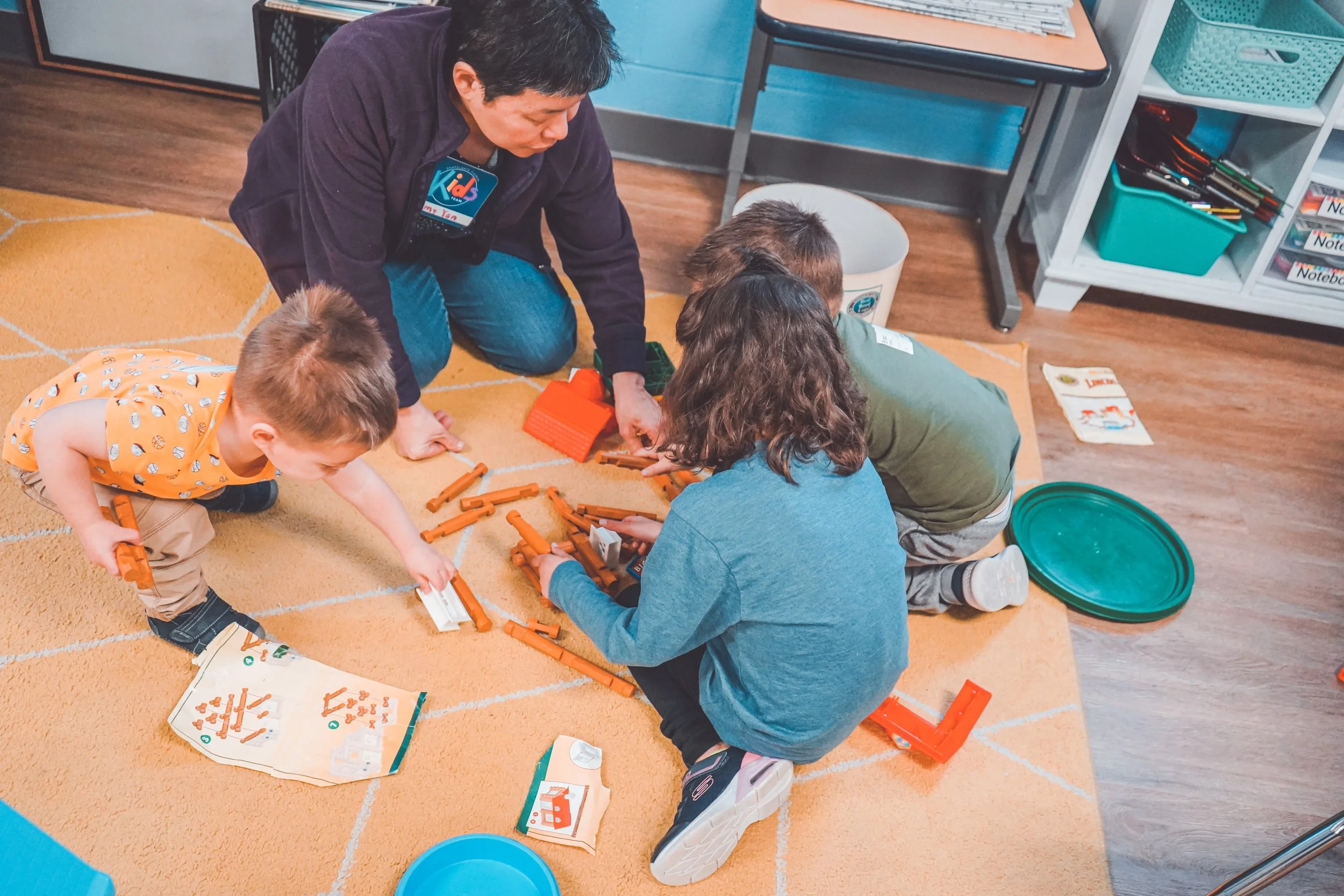 A teacher and four young children play with orange and white building blocks on an orange carpet in a classroom.