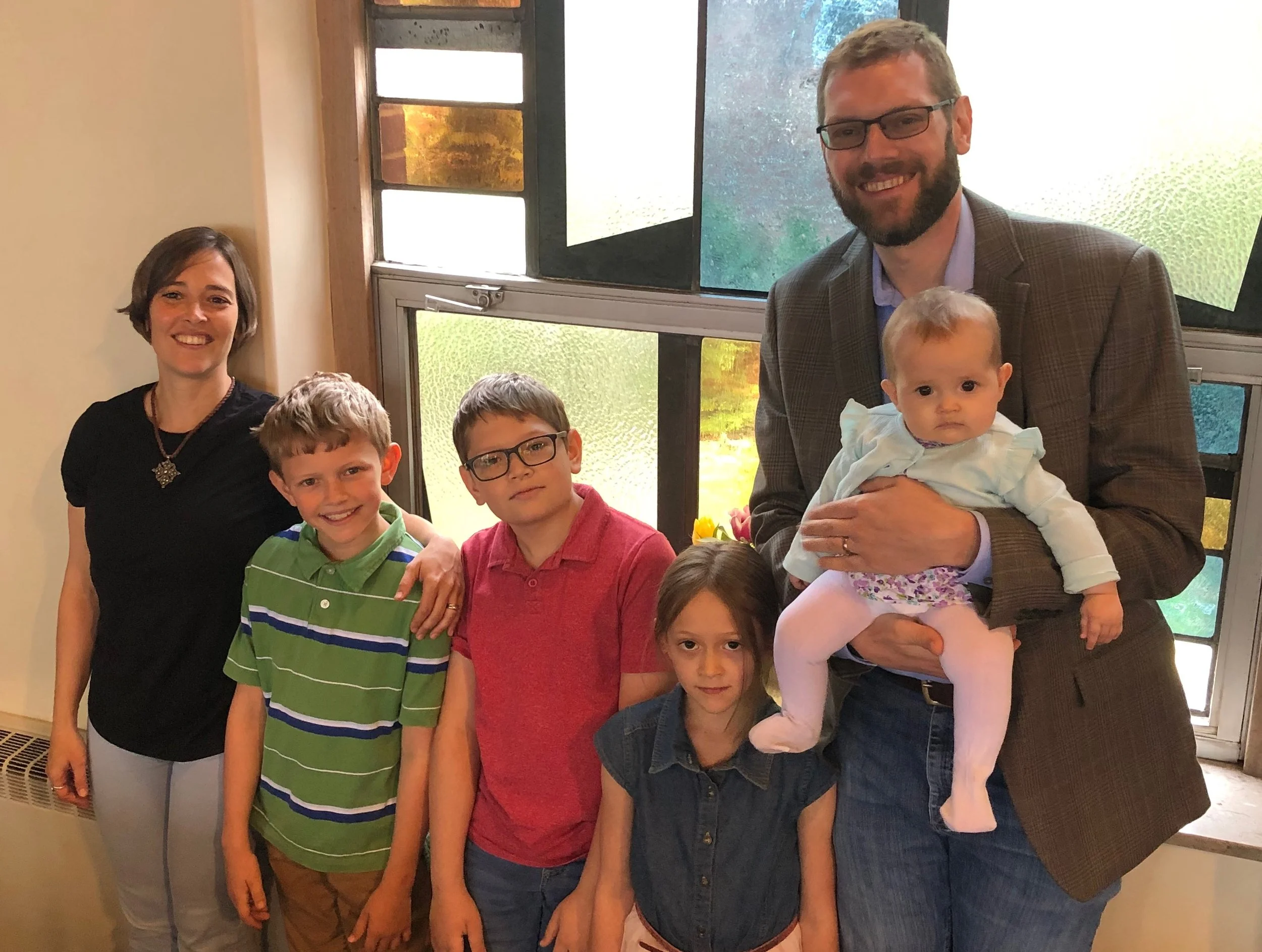 A group of six people, including five children and one adult man, are standing in front of a window. The man is holding a baby girl, and others are smiling for the photo.