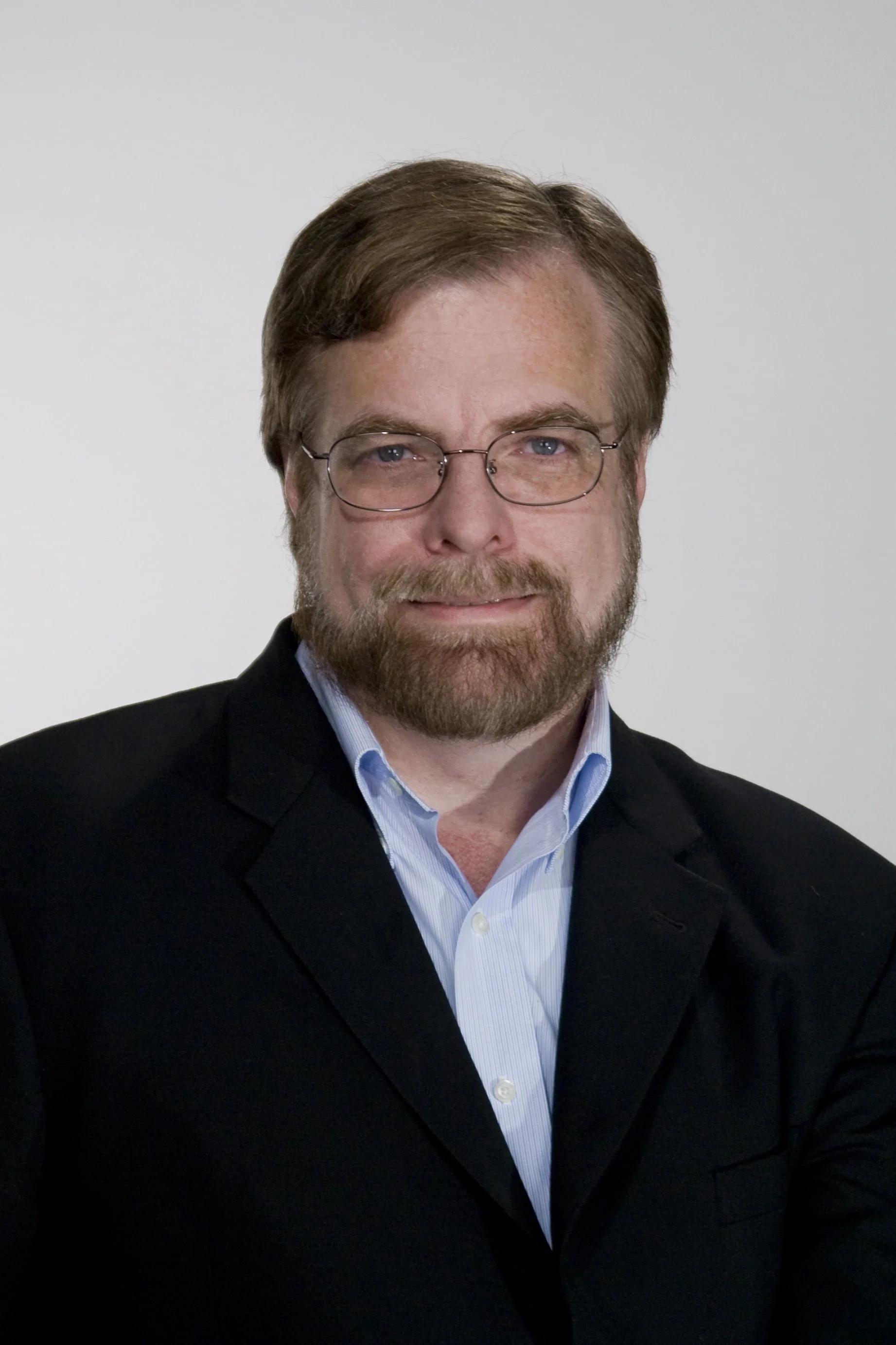 A middle-aged man with brown hair, glasses, and a beard, wearing a black blazer and light blue shirt, smiling at the camera against a plain white background.