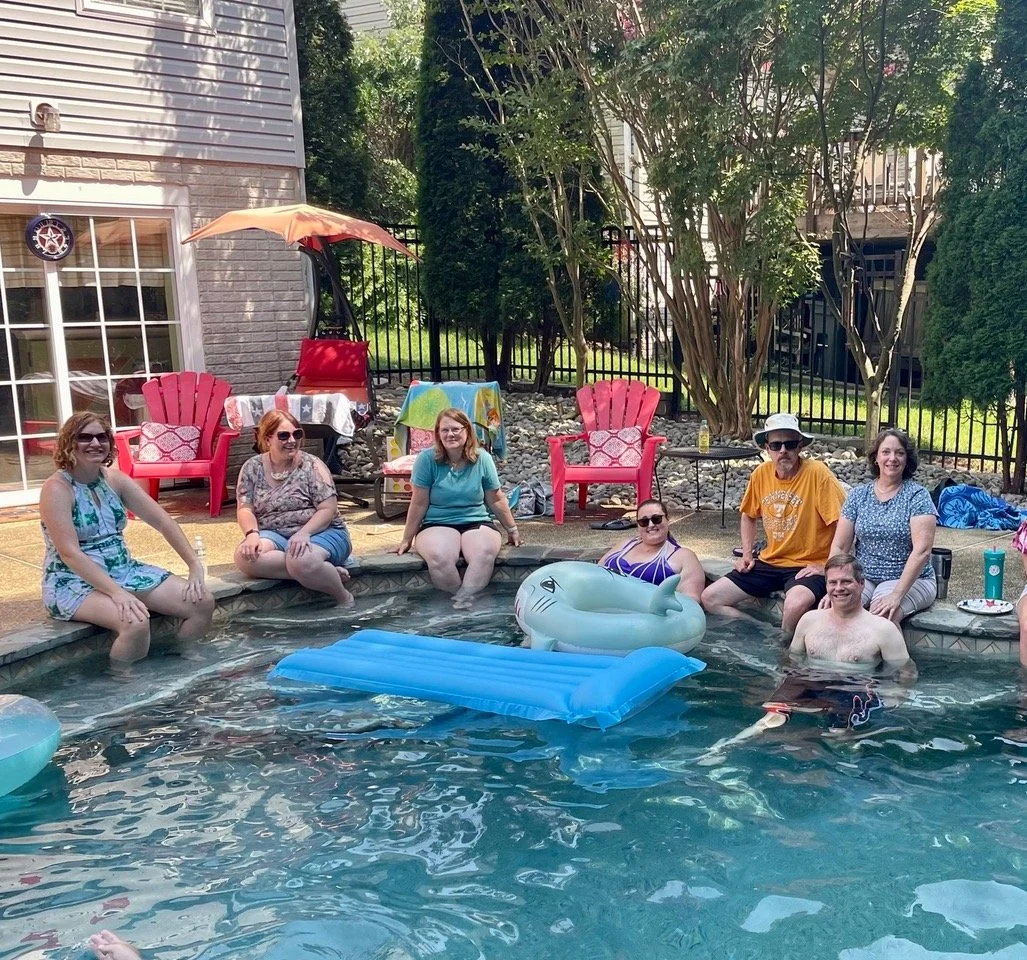 A group of people sitting on the edge of a backyard pool, smiling and relaxing with inflatable floats, surrounded by outdoor furniture and trees.