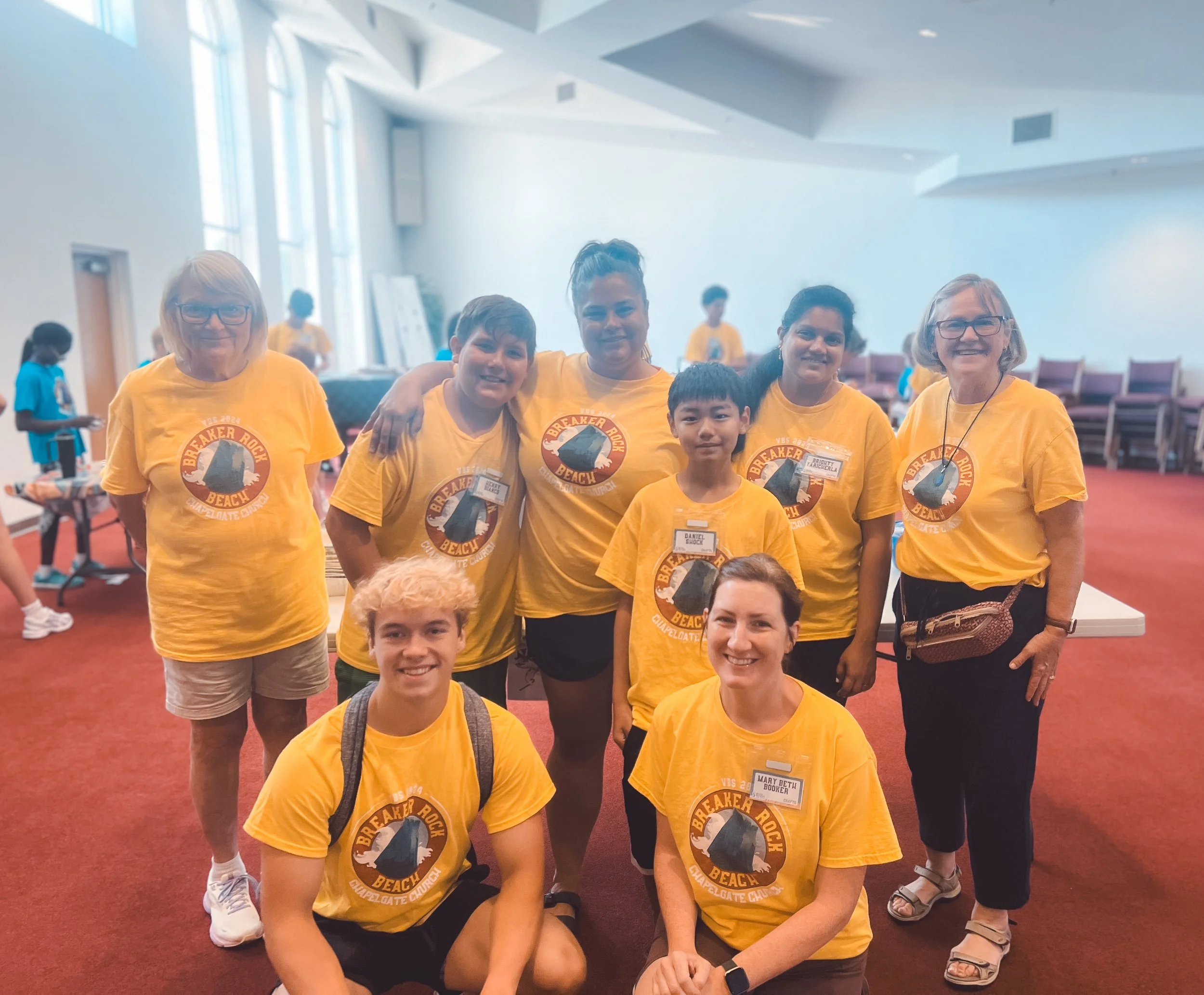 Group of people wearing yellow T-shirts with 'Breaker Rock Beach' logo, standing inside a large, well-lit room with red carpet.”}