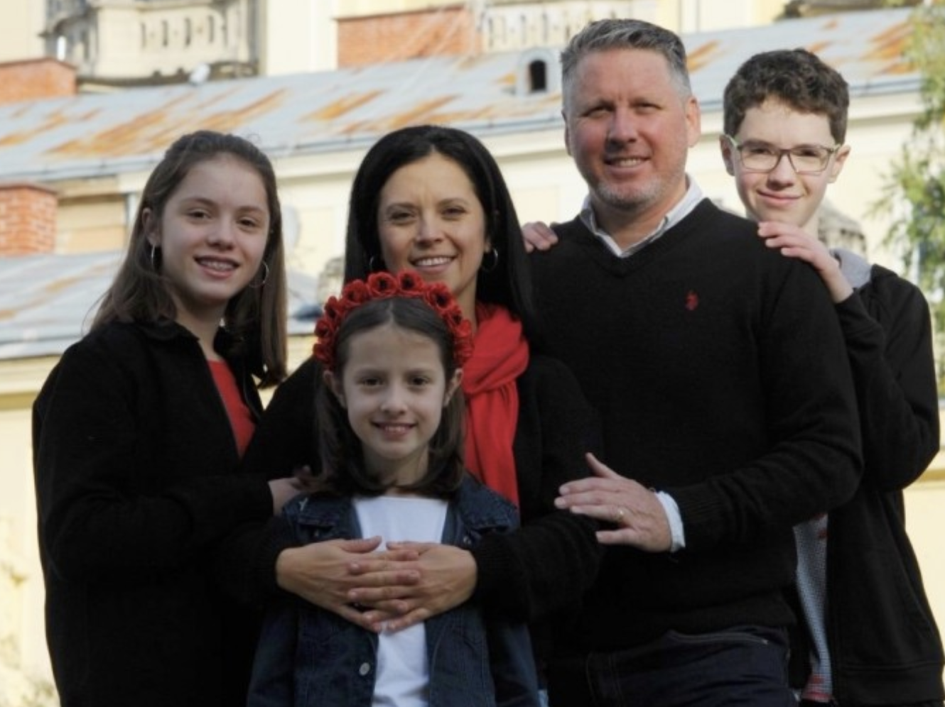 A family of six posing outdoors, smiling at the camera, with houses and trees in the background.