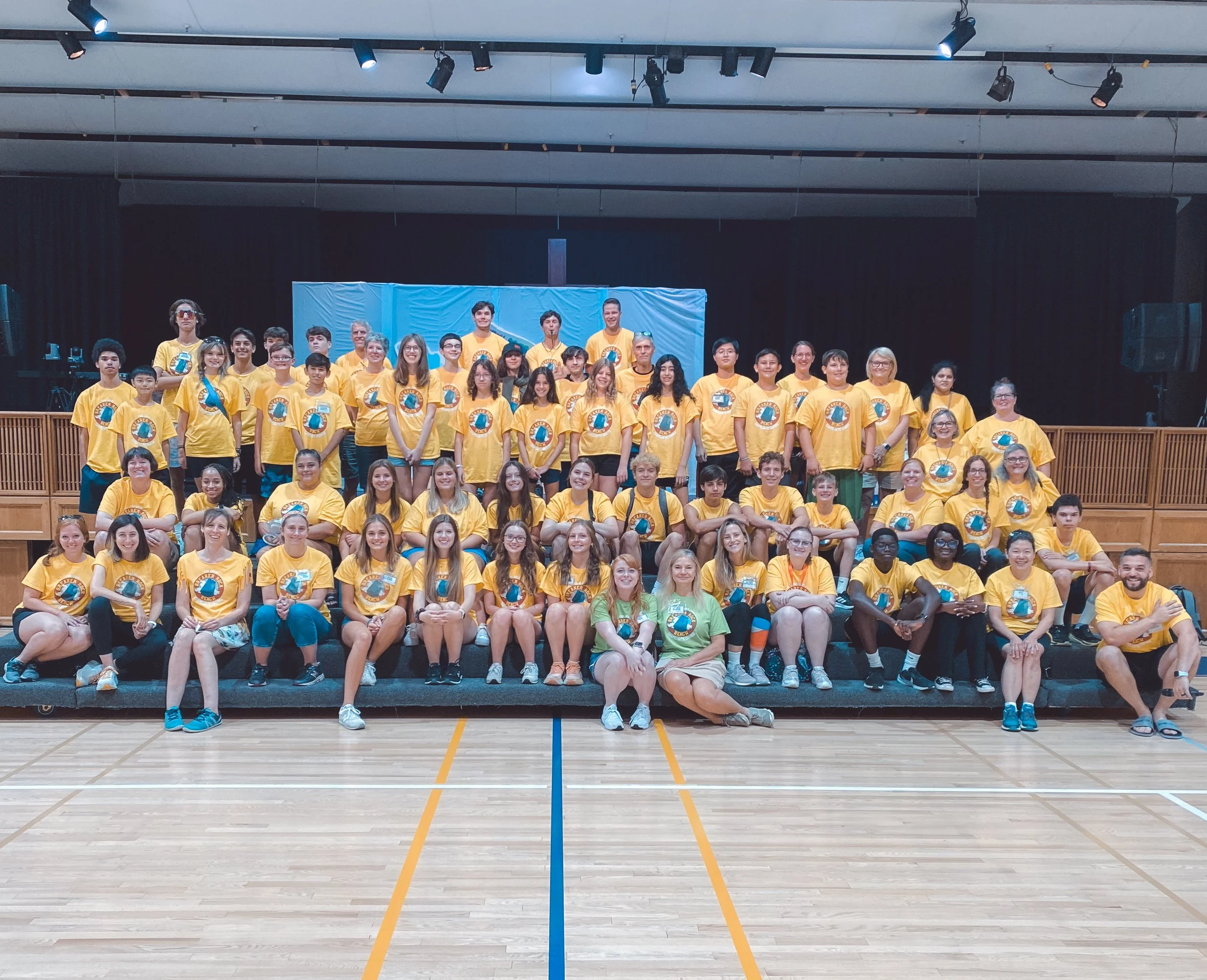 Group of young people and adult leaders wearing yellow T-shirts with a logo, posing for a group photo in an indoor gymnasium on a stage with a blue backdrop.