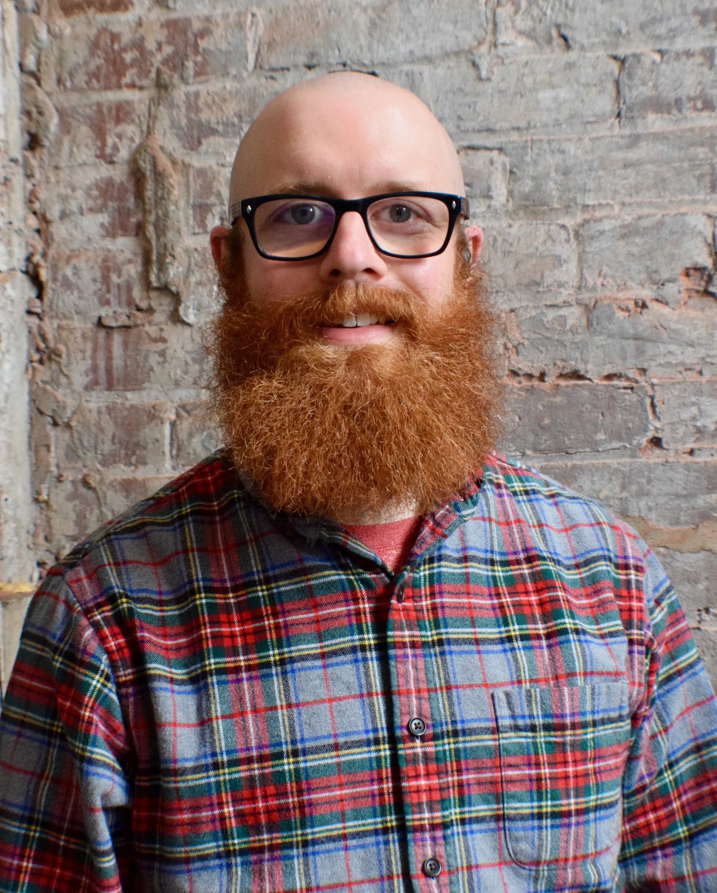A man with glasses and a red beard, wearing a plaid shirt, stands against a brick wall.