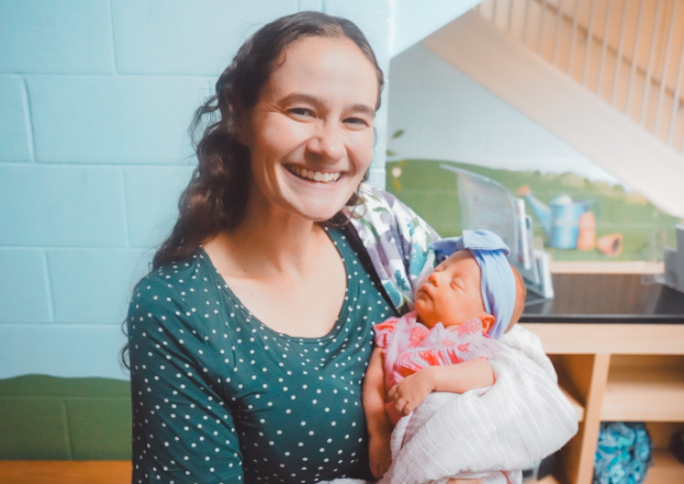 A woman with curly hair, glasses, and a dark patterned jacket smiling while holding a young boy in a classroom with colorful educational decorations and learning materials.