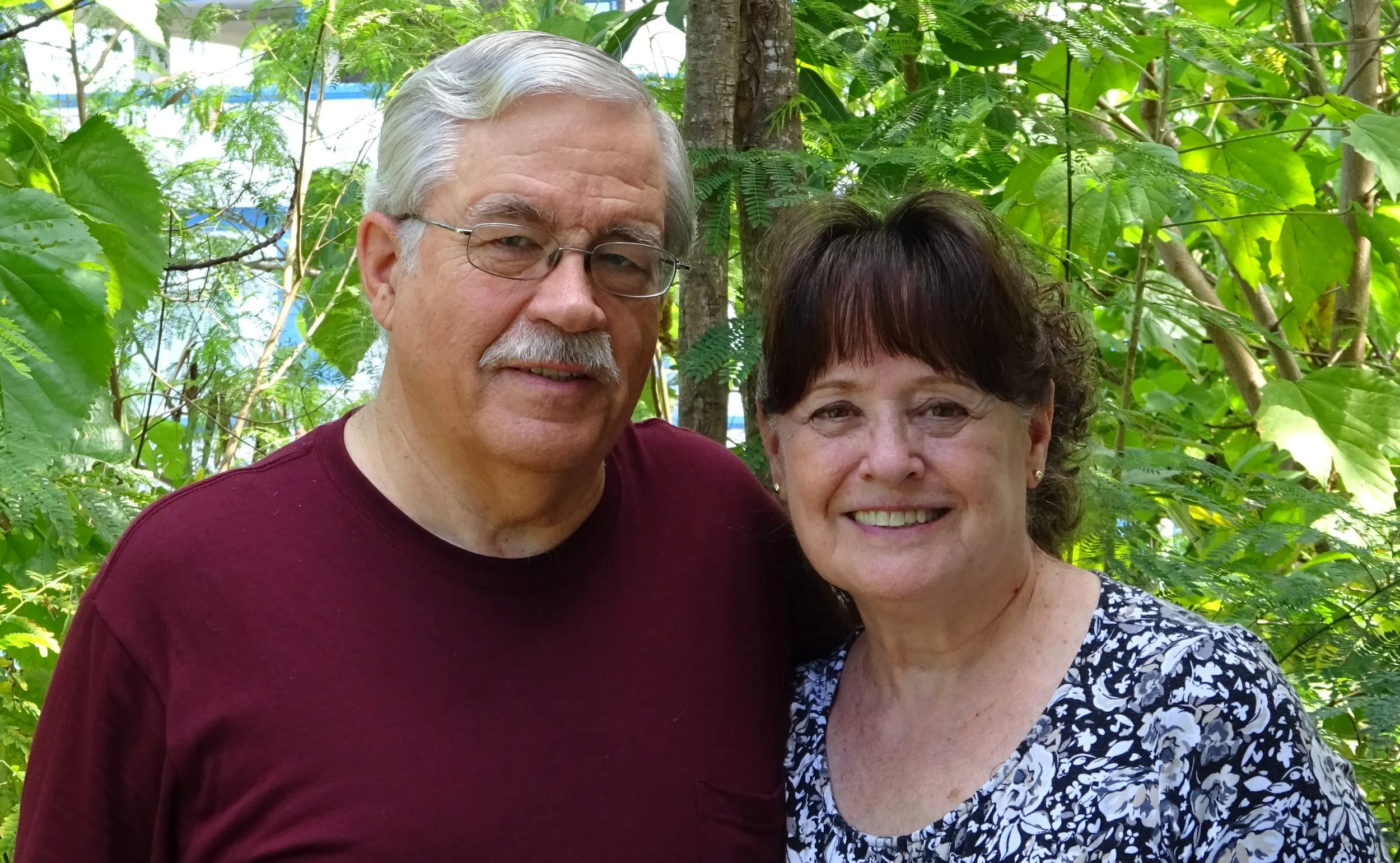 A man with gray hair, glasses, and a mustache, and a woman with short dark hair, smiling, standing outdoors among green leafy trees.