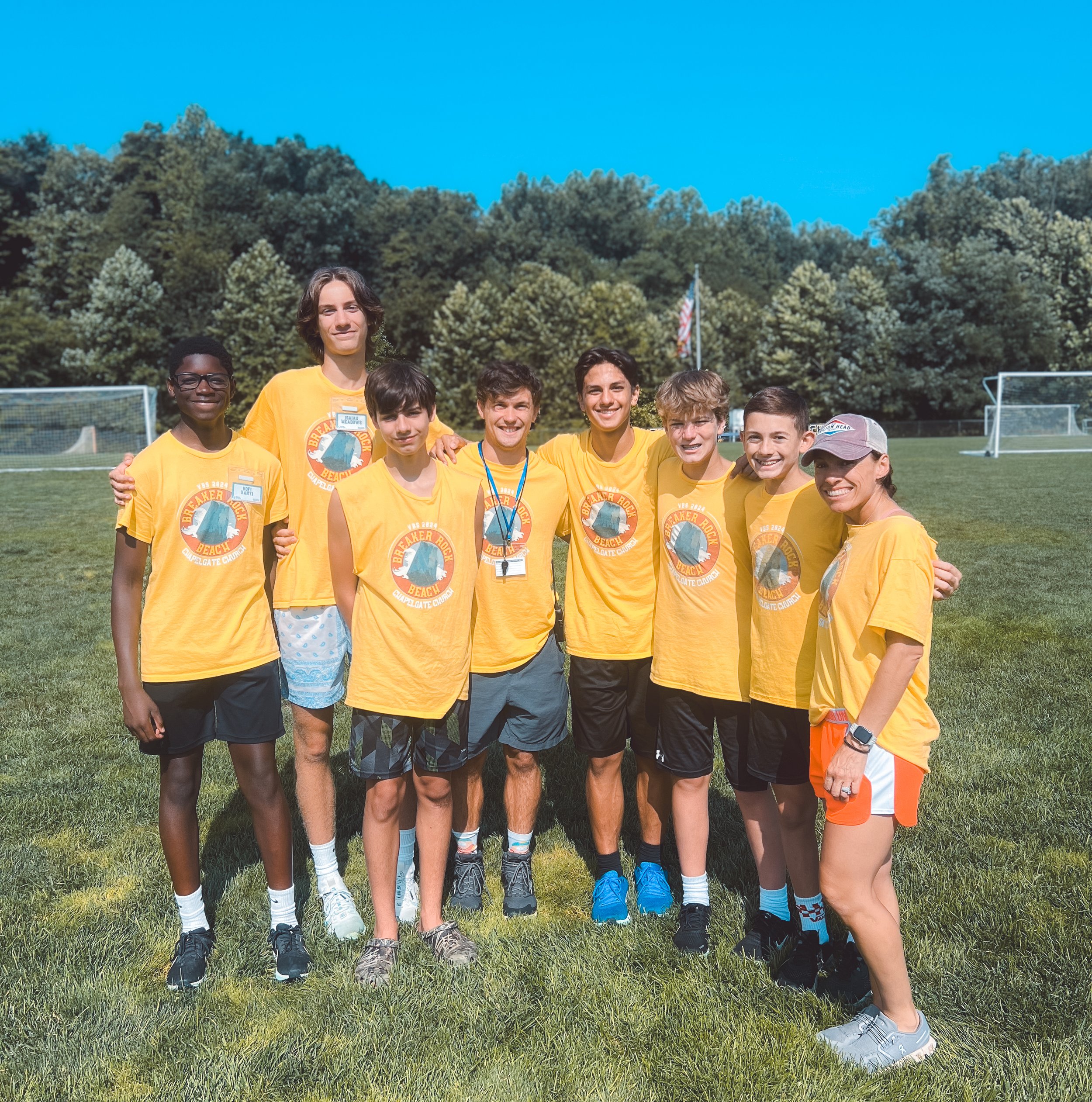 Group of young people wearing yellow T-shirts standing on a grassy field with soccer goals and trees in the background.