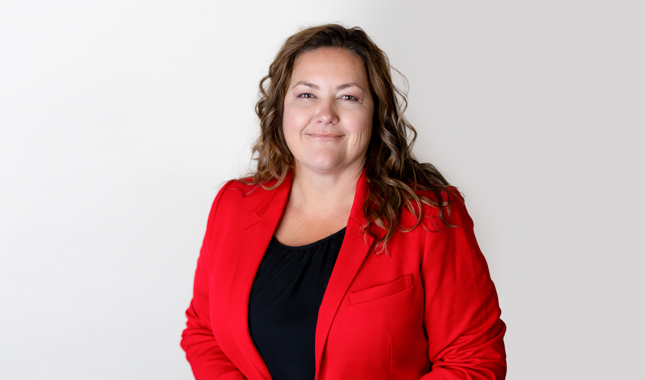 A woman with curly brown hair wearing a red blazer over a black top, smiling, against a plain white background.