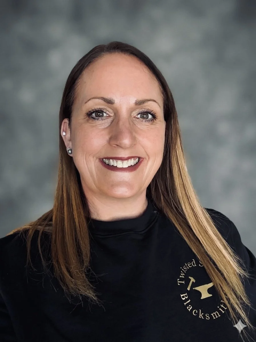 A woman with long brown hair, wearing earrings, smiling, and wearing a black shirt with gold-colored text and logo on the chest, against a gray background.