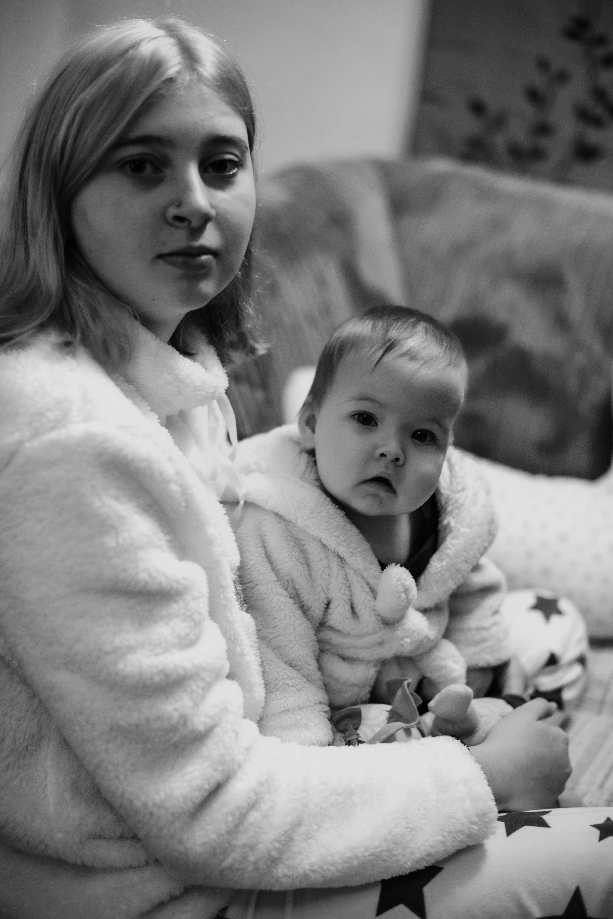 A woman and a young girl sitting close together, both wearing cozy, fluffy jackets, with the woman looking at the camera and the girl gazing downward, in a casual indoor setting.