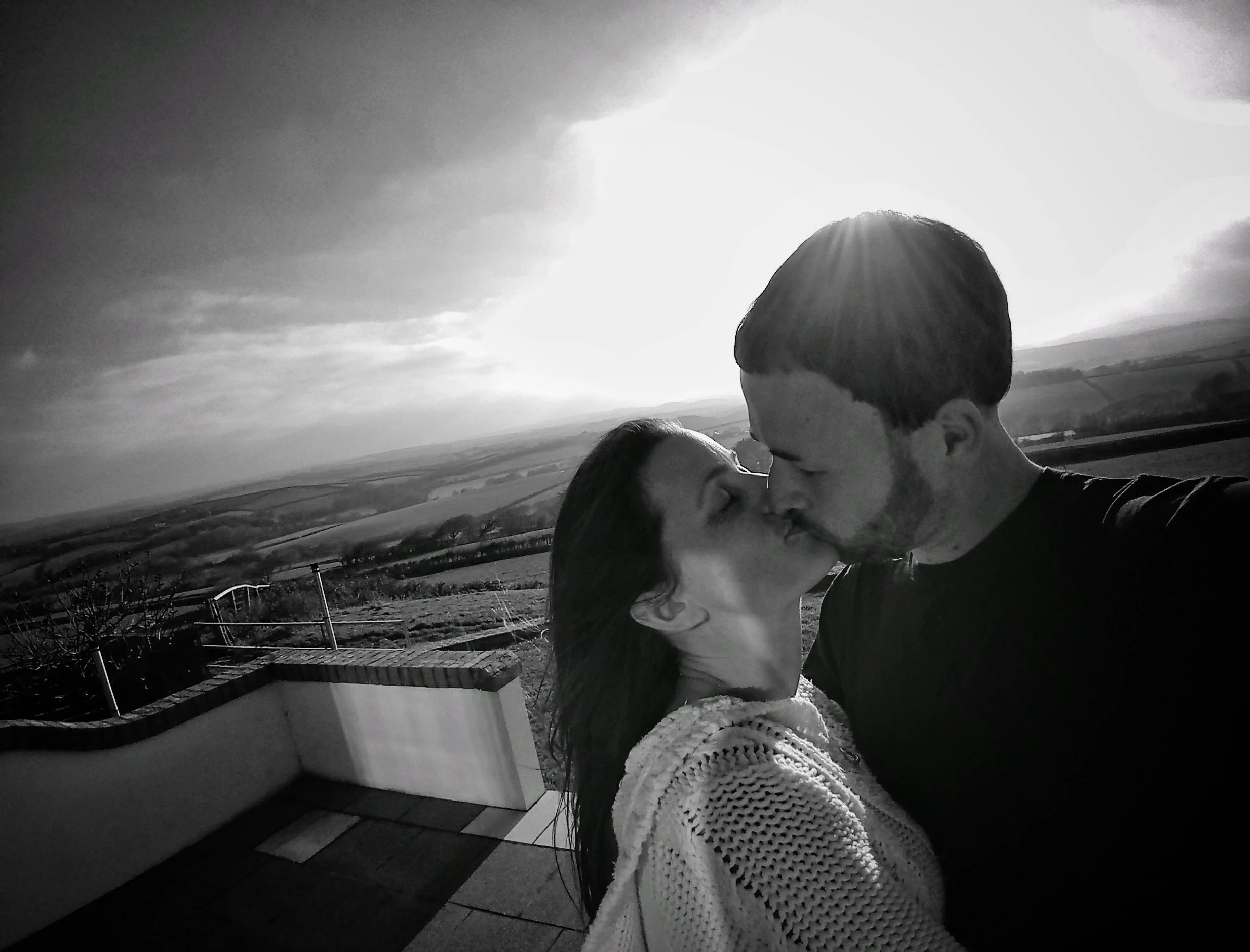 A couple sharing a kiss outdoors with a scenic rural landscape in the background, backlit by bright sunlight.