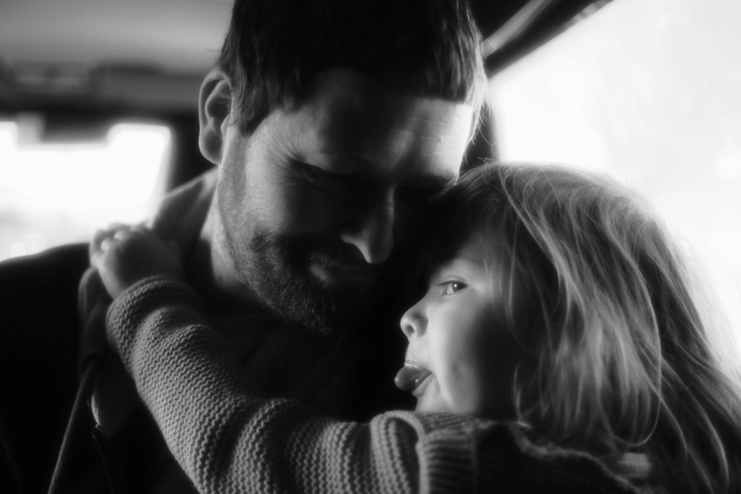 A black and white photograph of a man with a beard and a young girl. The man is resting his forehead against the girl's, and they are closely embracing. The girl has light hair and is looking upwards.