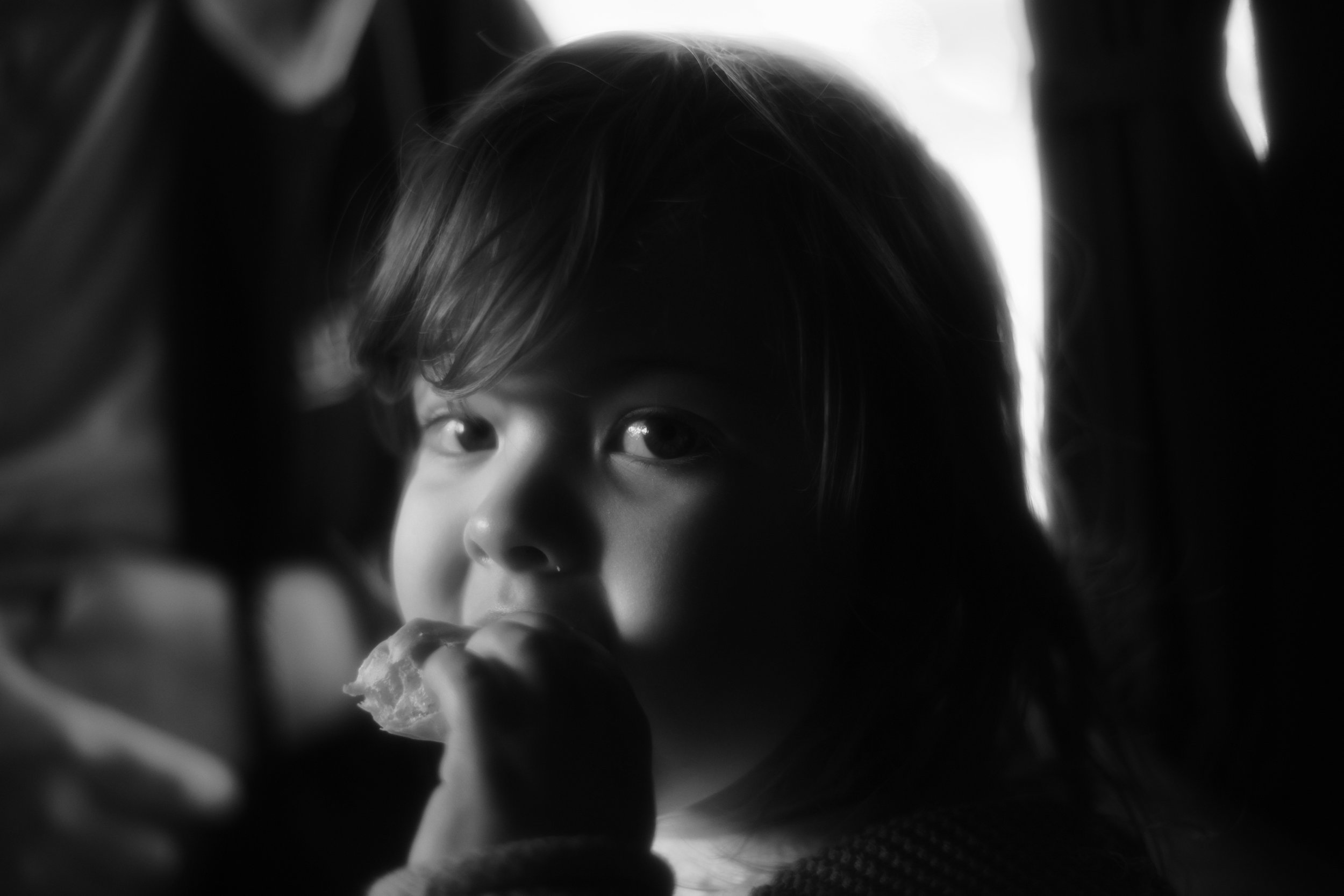 A child with short hair and wide eyes, holding a piece of food near their mouth, viewed through a dark, shadowed opening.