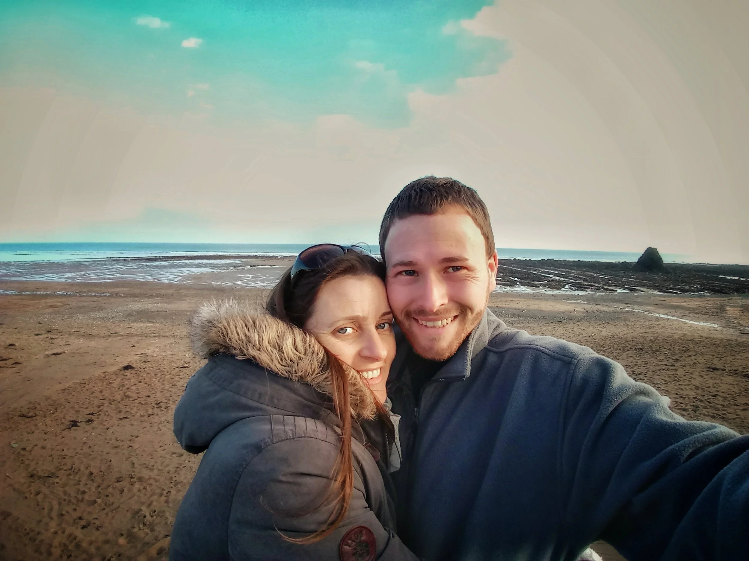 A smiling couple taking a selfie on a beach with a muddy shoreline, calm ocean, and cloudy sky in the background.