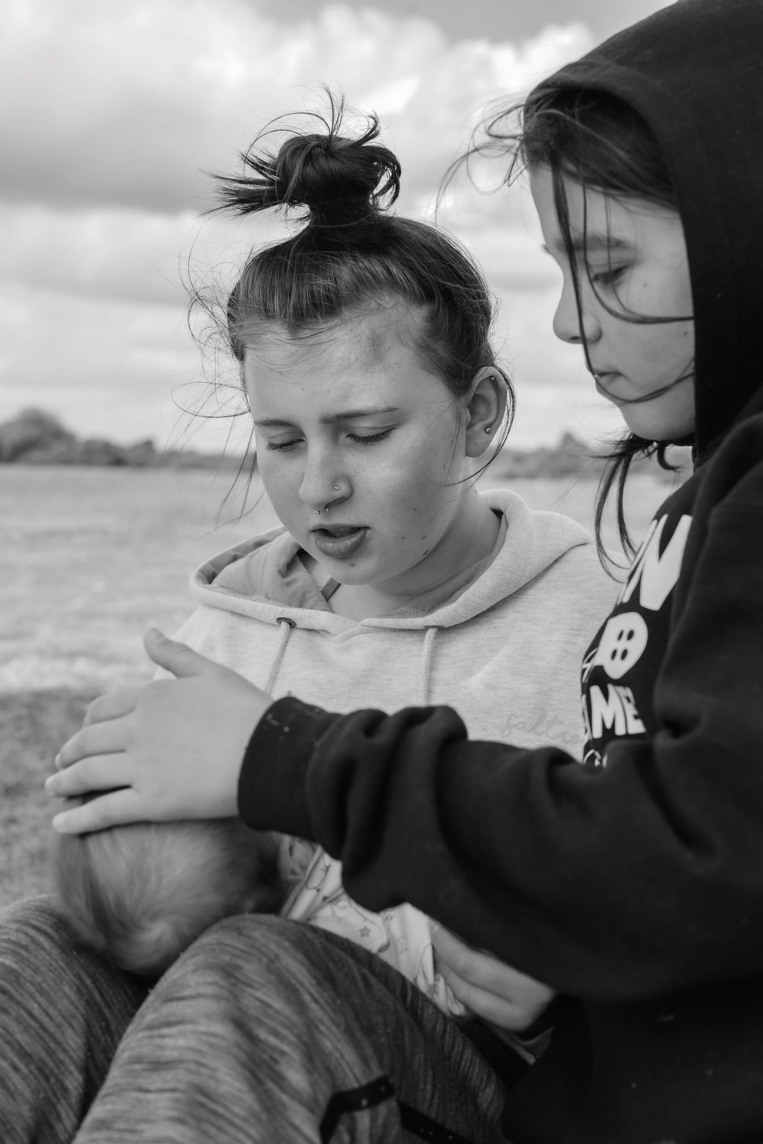 Two young women sitting outdoors, looking at a smartphone together. One wearing a hoodie, the other with a top knot hairstyle, both focused on the phone. Overcast sky in background.