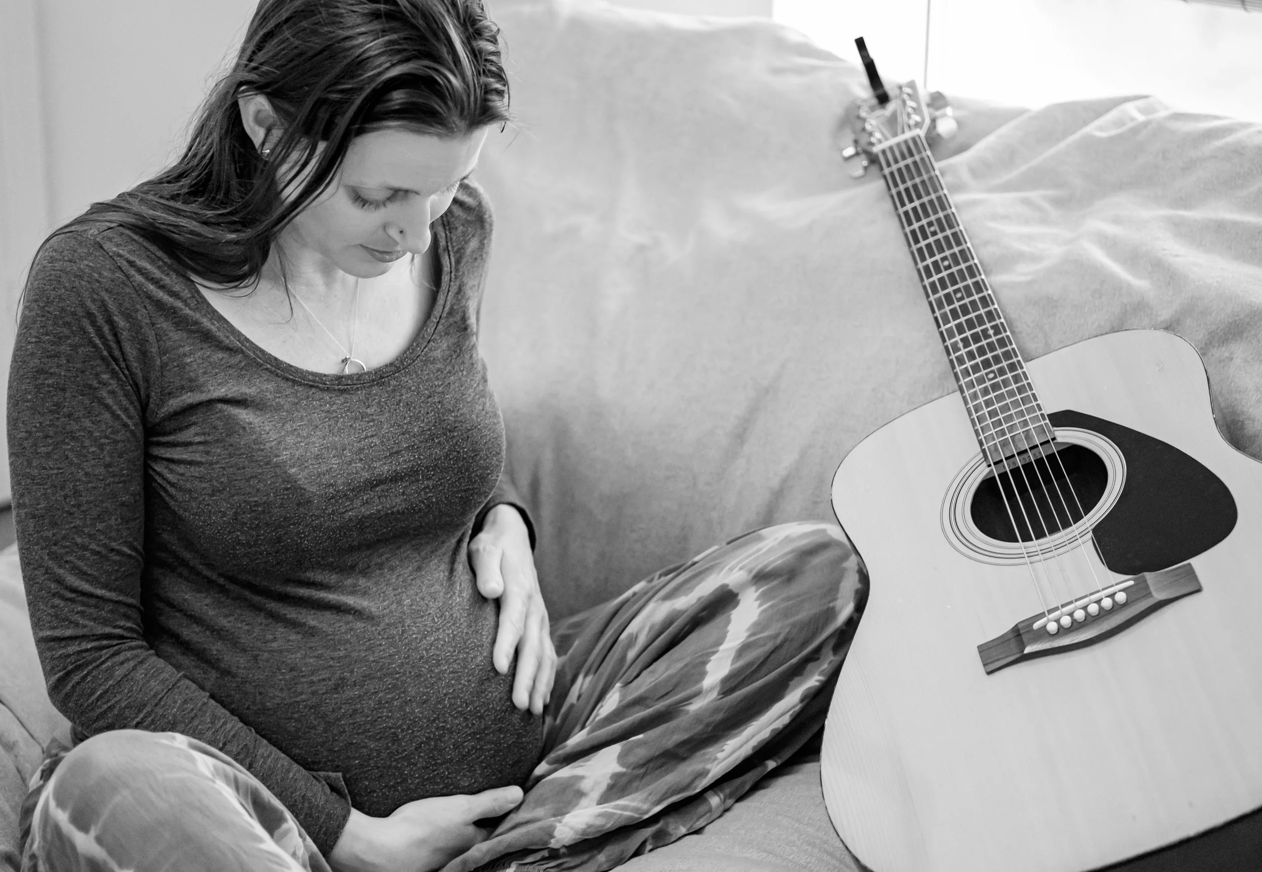 A young woman with long dark hair, wearing a long-sleeve shirt, sitting on a floor with her right hand on her stomach, looking down. Next to her is an acoustic guitar resting against a surface, with a black strap hanging from it. The background featu