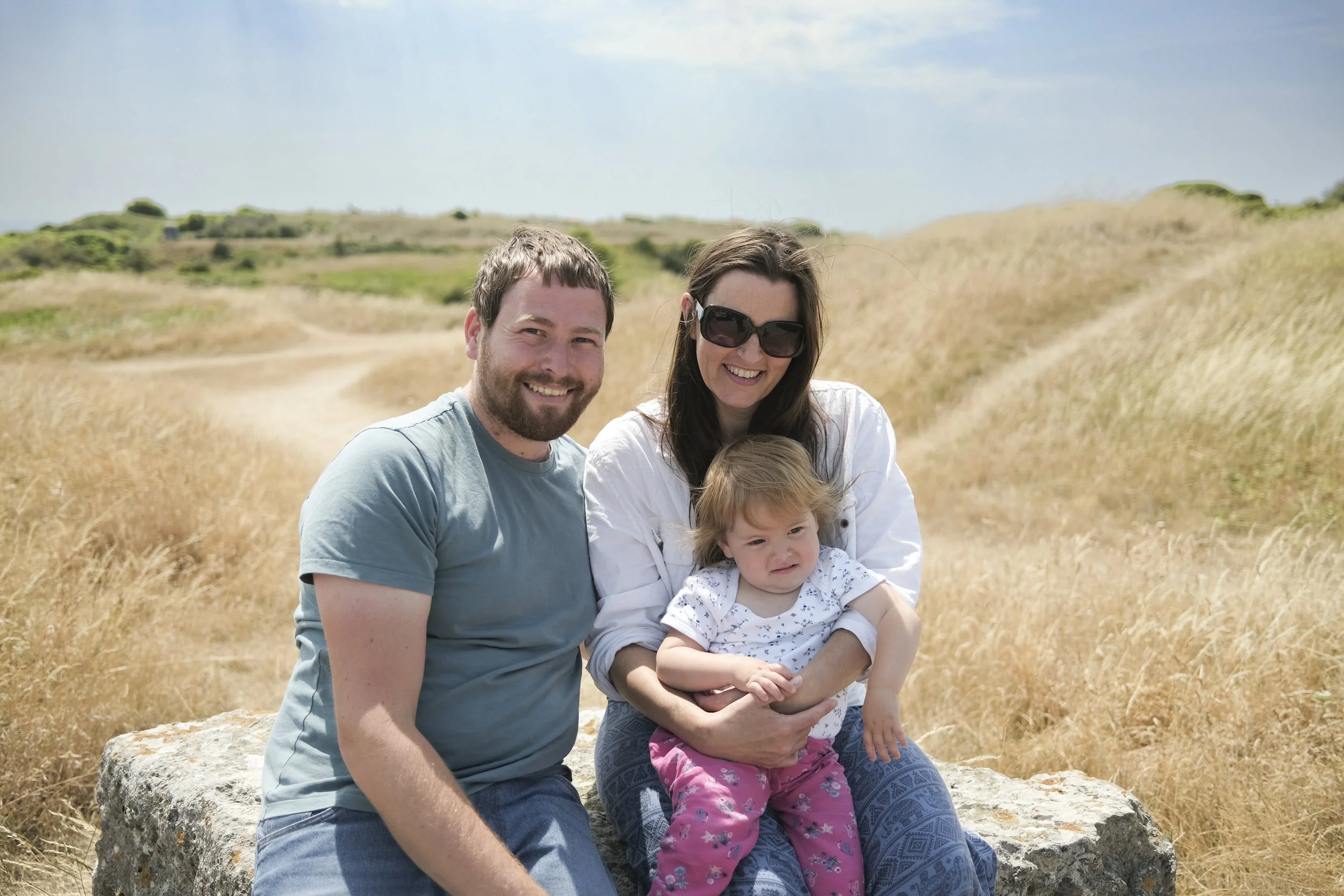 A family, including a man, woman, and young girl, sitting on a large rock in a field of tall, dry grass under a partly cloudy sky.