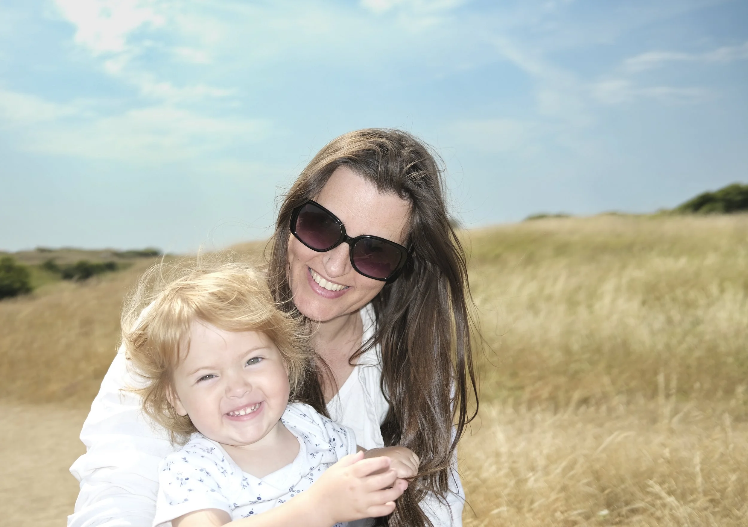 A woman with long brown hair wearing sunglasses smiling and holding a young girl with curly blonde hair and a white shirt, outdoors in a grassy field with a blue sky and some clouds.