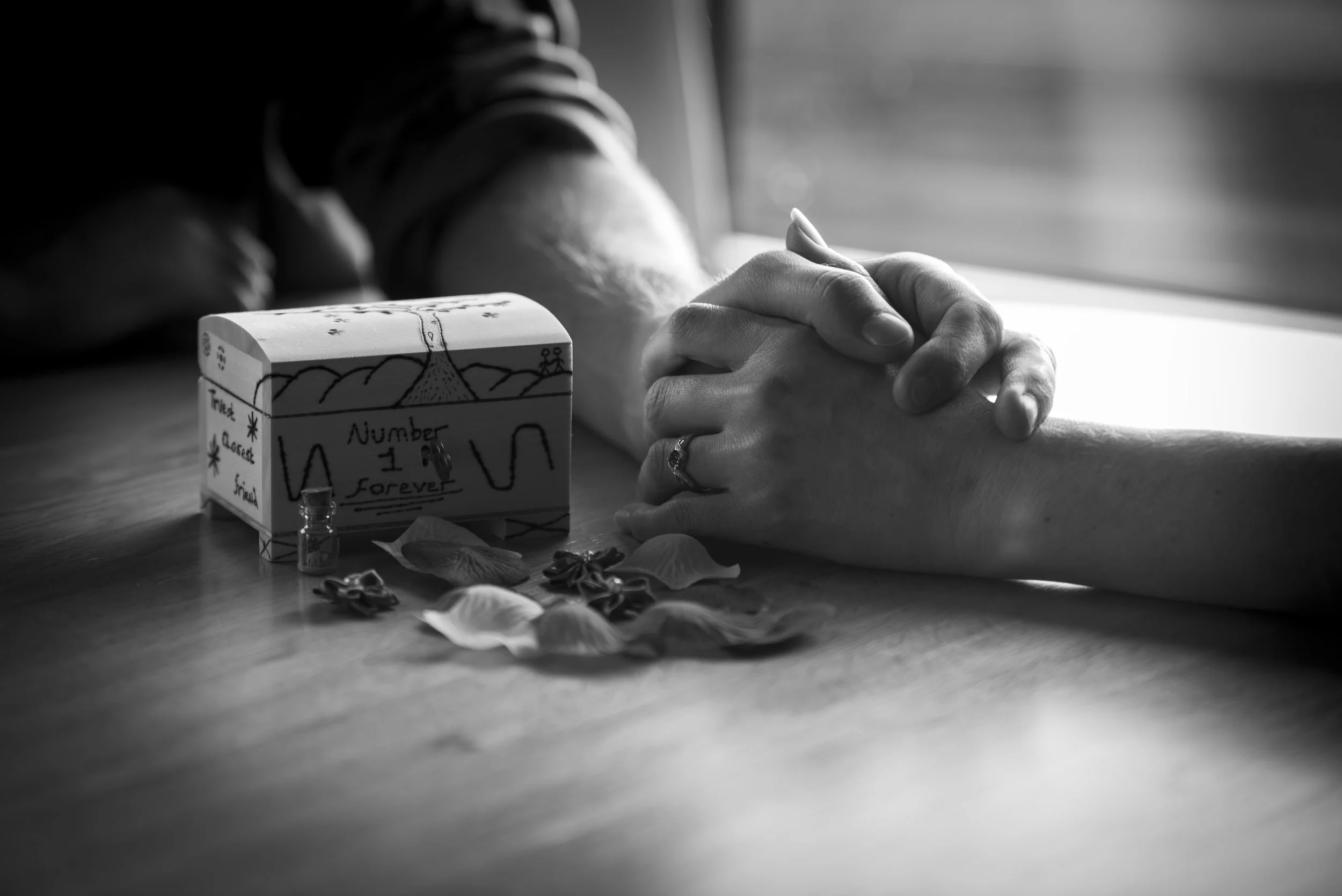 Close-up of two people holding hands on a table, with a decorated box, small bottles, flower petals, and other objects in black and white.