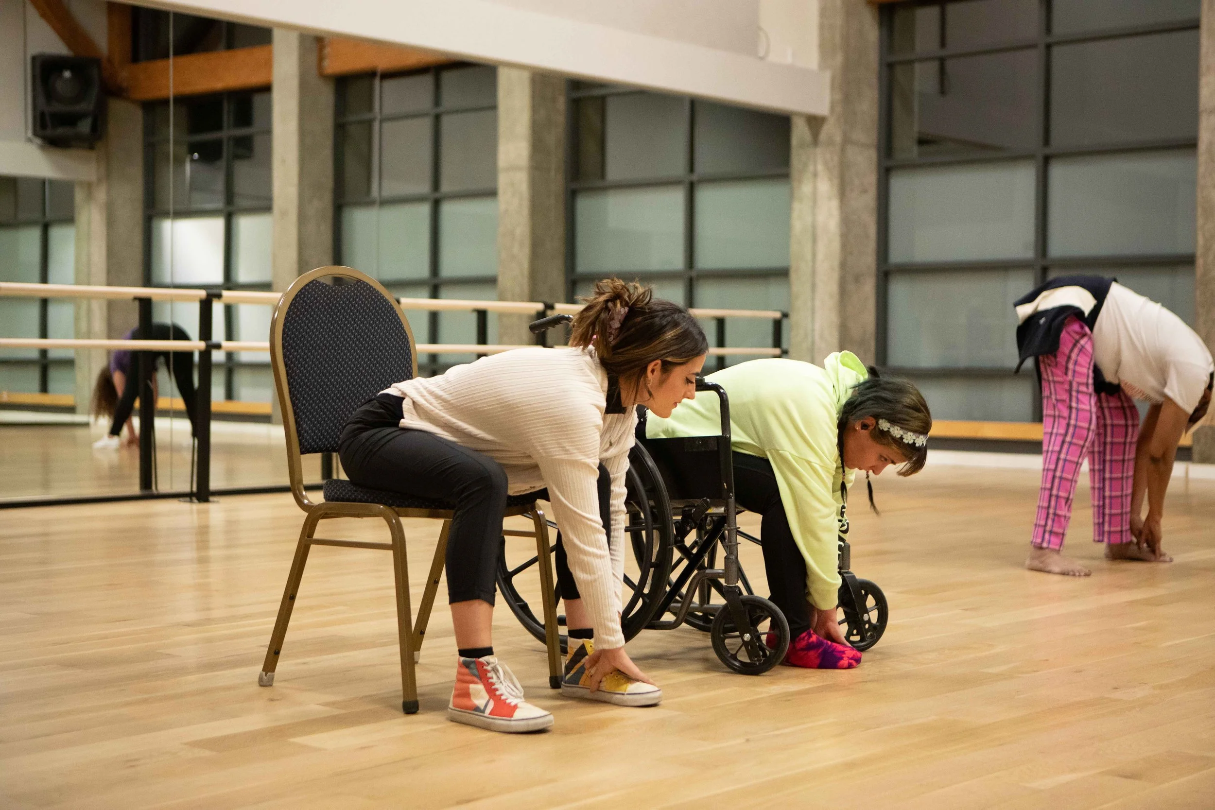 People practicing yoga or stretching in a dance studio. The room has wooden floors, mirrors, and ballet barres.