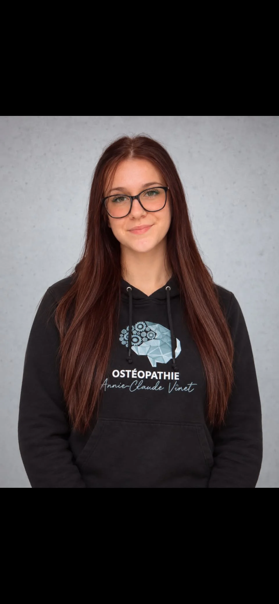 Jeune femme avec lunettes, cheveux longs roux, portant un sweat noir avec le logo d'une tête de cerveau et le texte 'OSTÉOPATHIE Annie-Claude Veret', posant devant un mur gris.