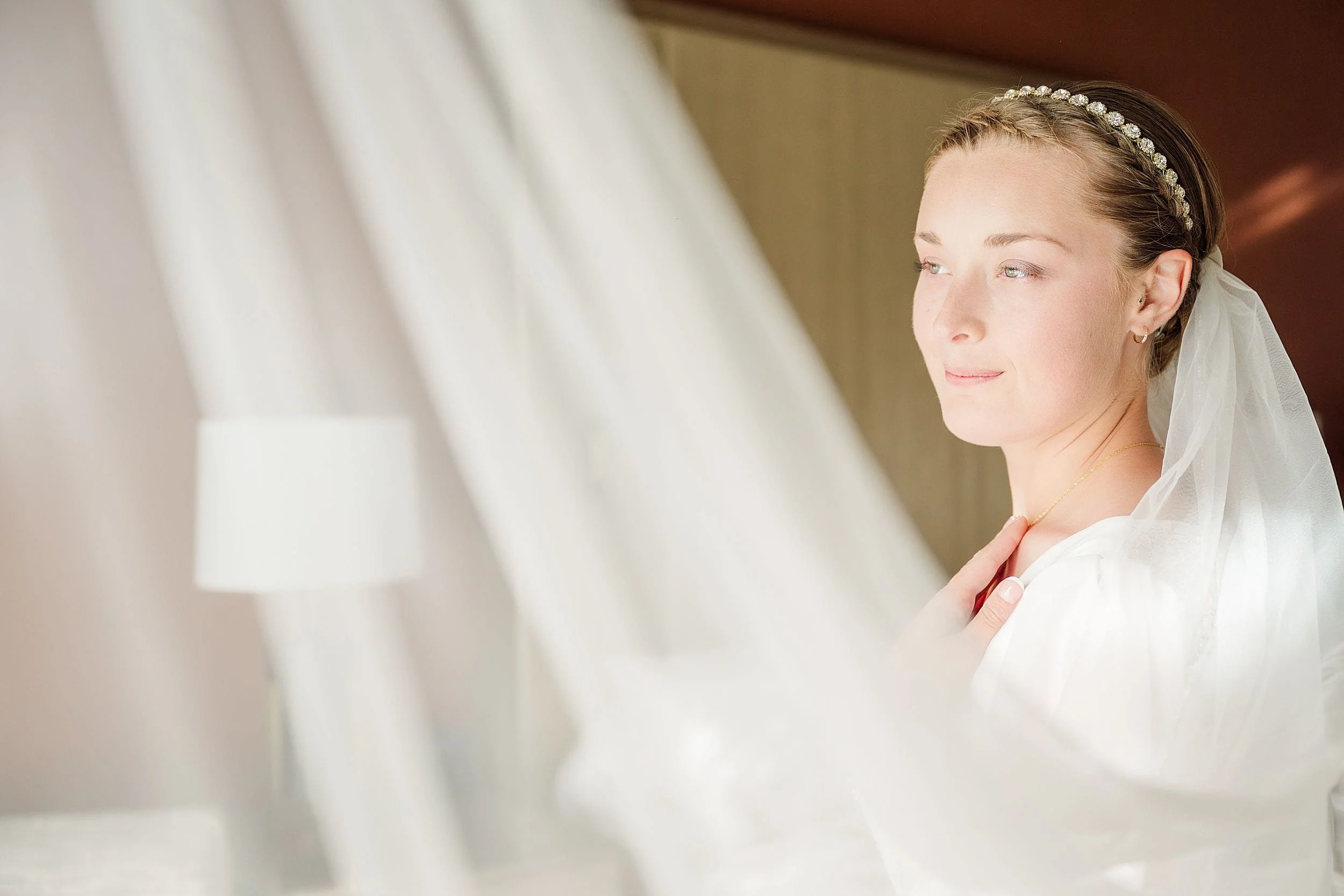 Bride in white dress with sheer veil, wearing a pearl headband, gazes softly to the side, emitting a serene and contemplative mood.