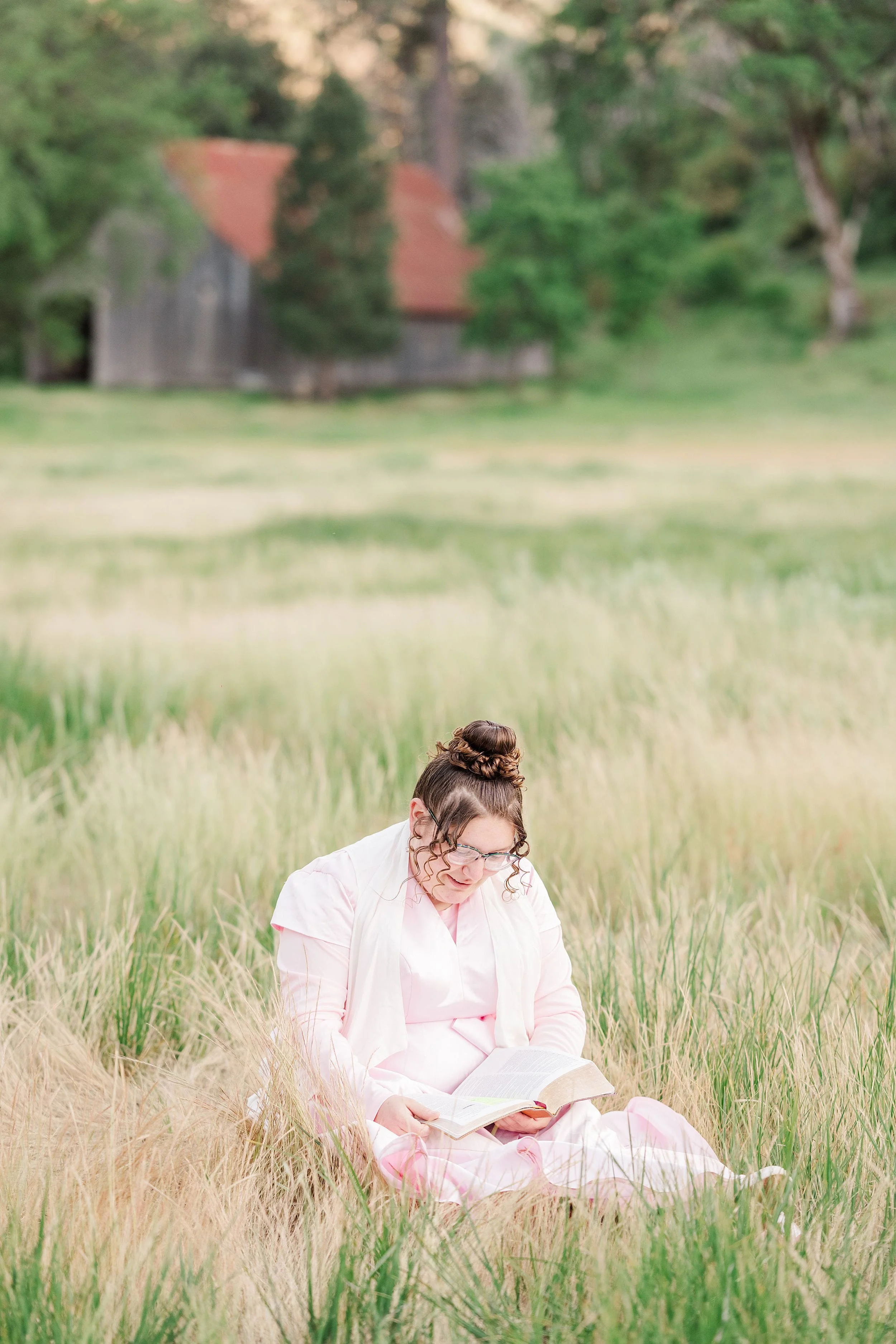 A person in a pink dress sits in a field of tall grass, reading the bible. Behind them, a rustic barn with a red roof is surrounded by lush greenery.