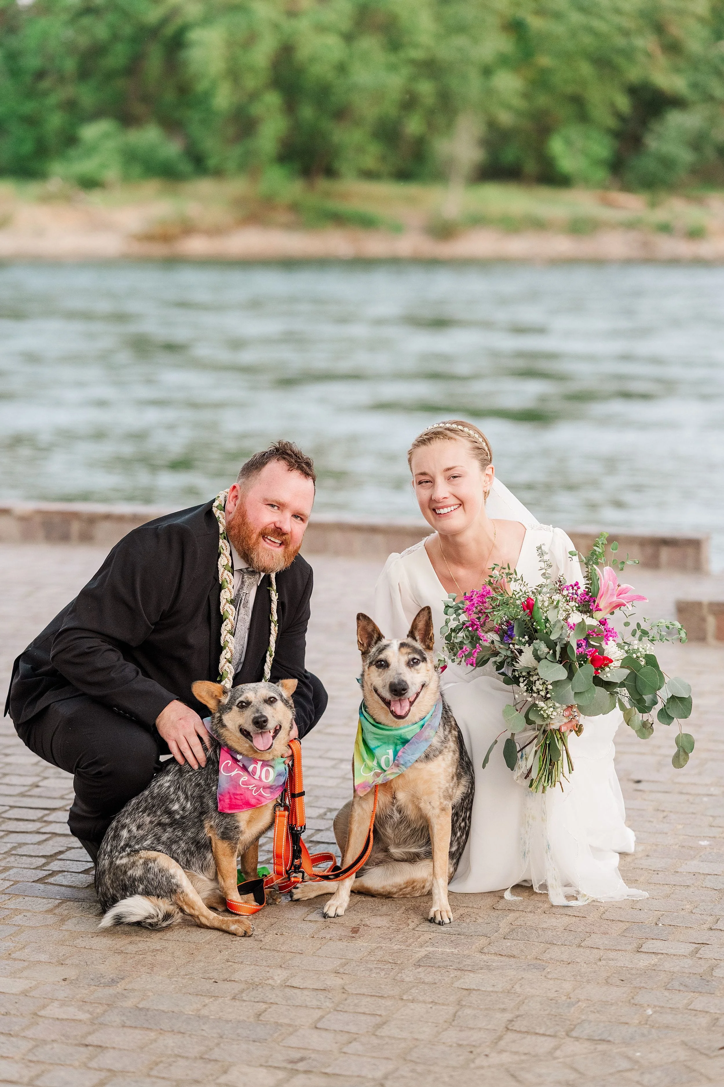 A couple in wedding attire smiles beside two dogs with colorful bandanas, posing by a serene river, conveying joy and celebration.