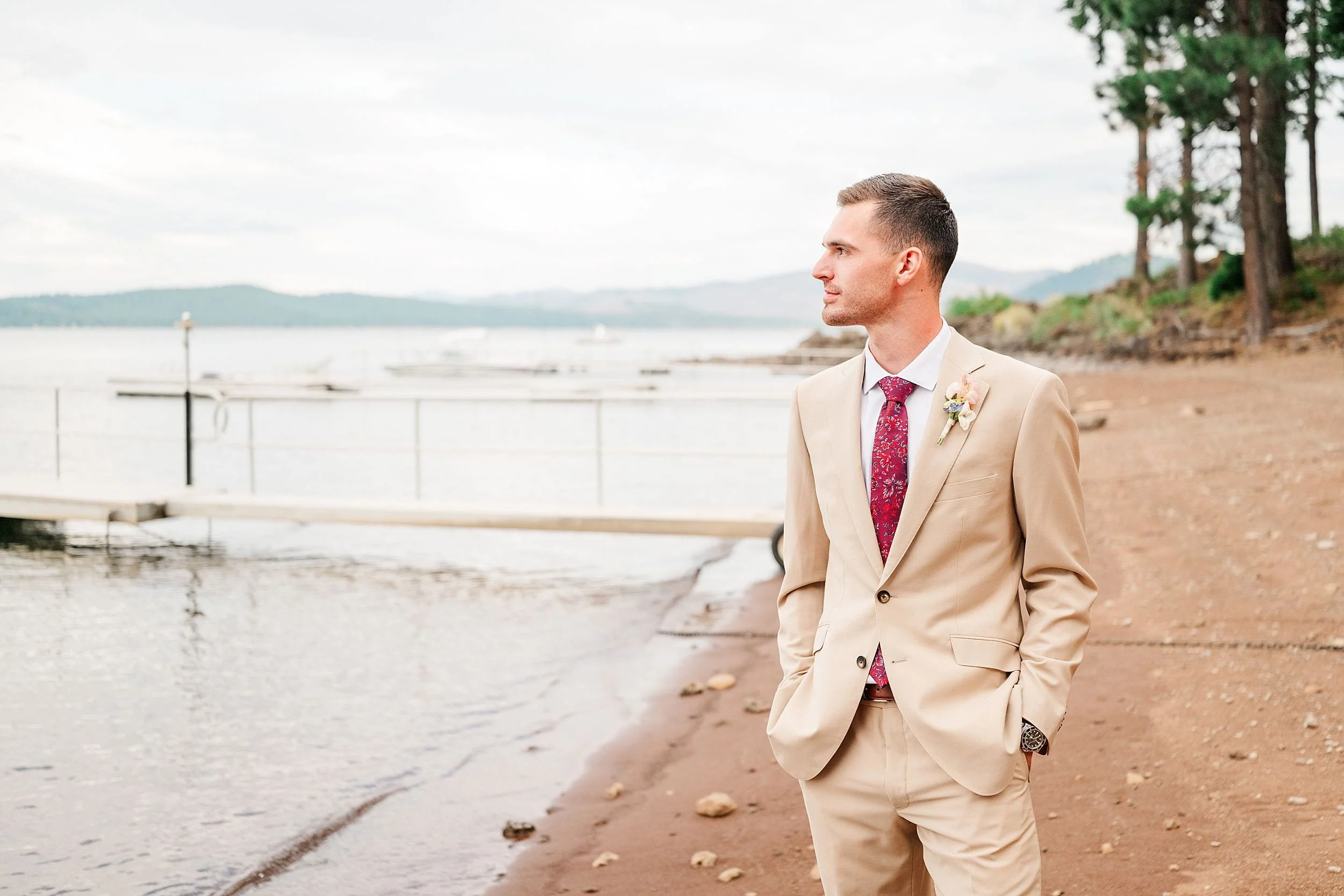 Groom in a tan suit, red tie, and boutonniere stands thoughtfully on a lakeshore, with a dock, trees, and hazy hills behind him.