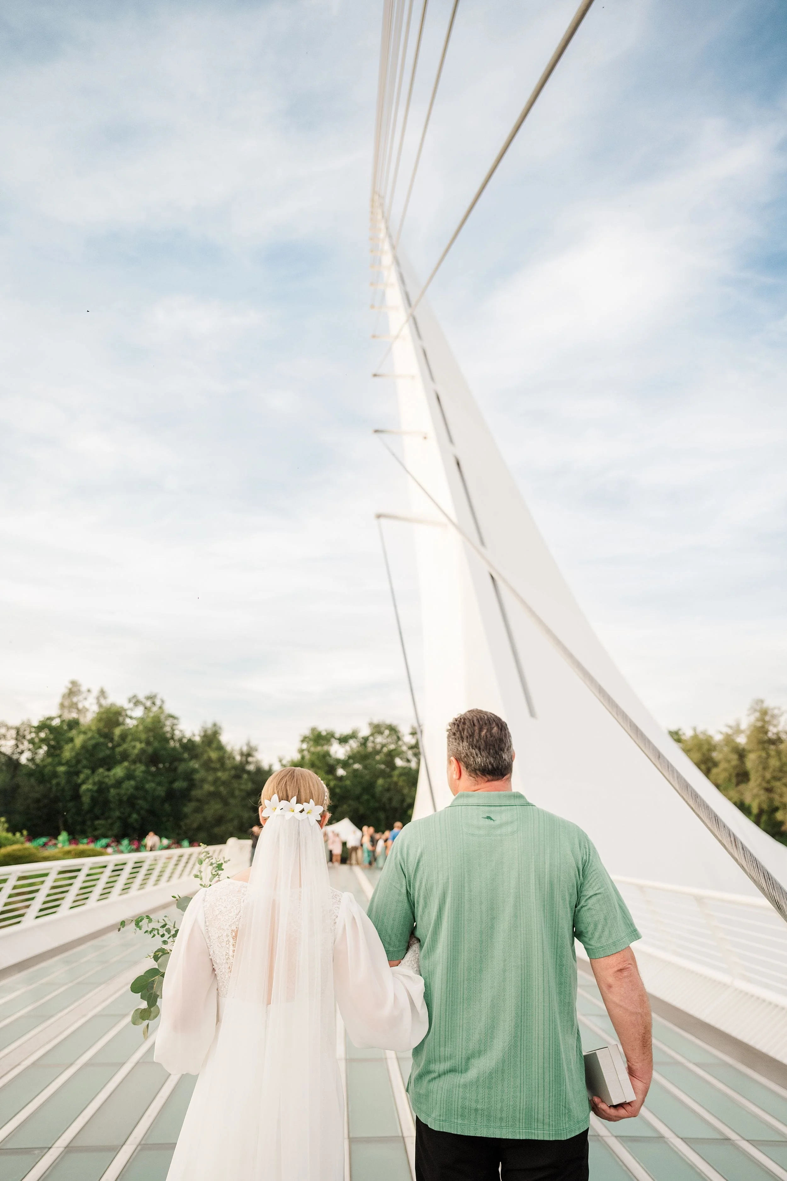 Bride in white gown and veil walks arm-in-arm with a man in a green shirt across a modern suspension bridge, under a clear blue sky.