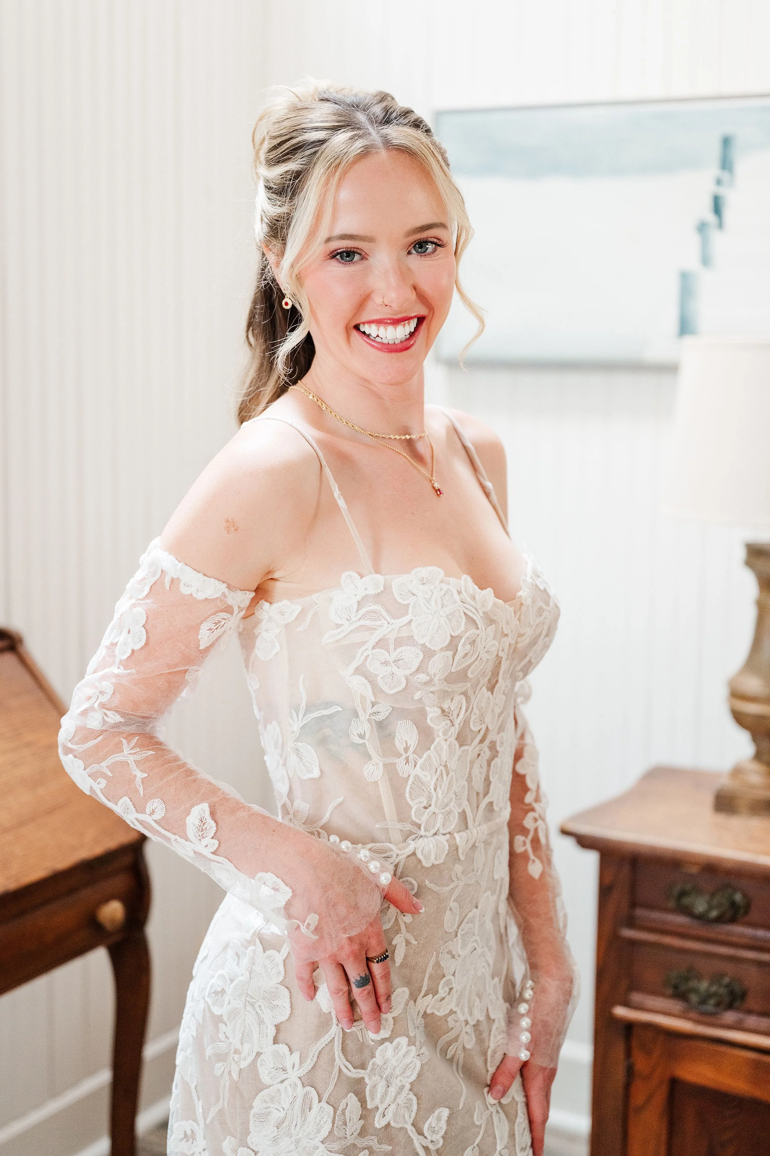 Smiling bride in an off-the-shoulder white lace wedding dress poses in an elegant room, conveying joy and bridal elegance.