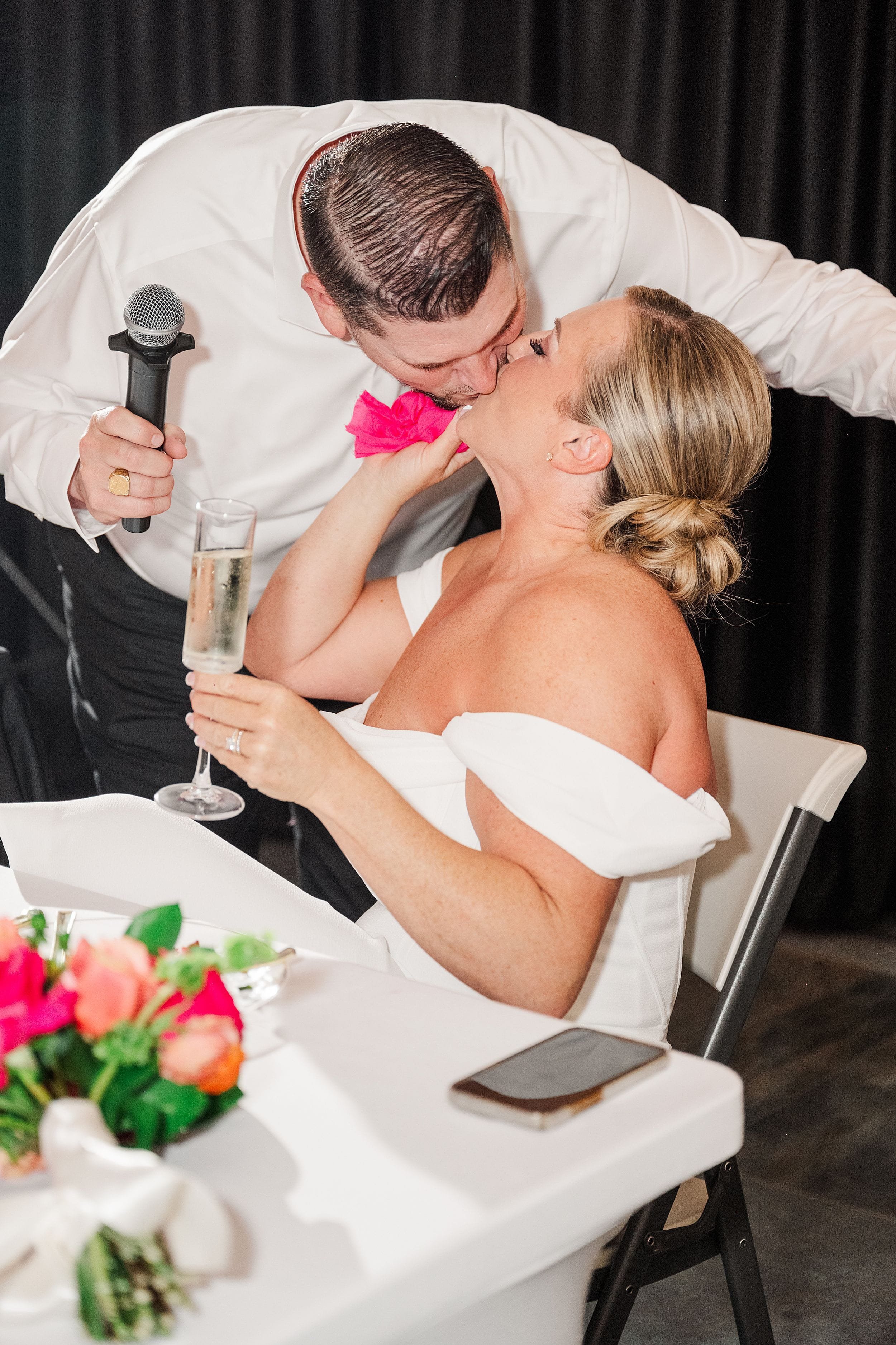 A man in a white shirt holds a microphone, leaning over to kiss a woman in an off-shoulder white dress. She smiles with a champagne glass. Vibrant bouquet and phone on table suggest a celebratory event.