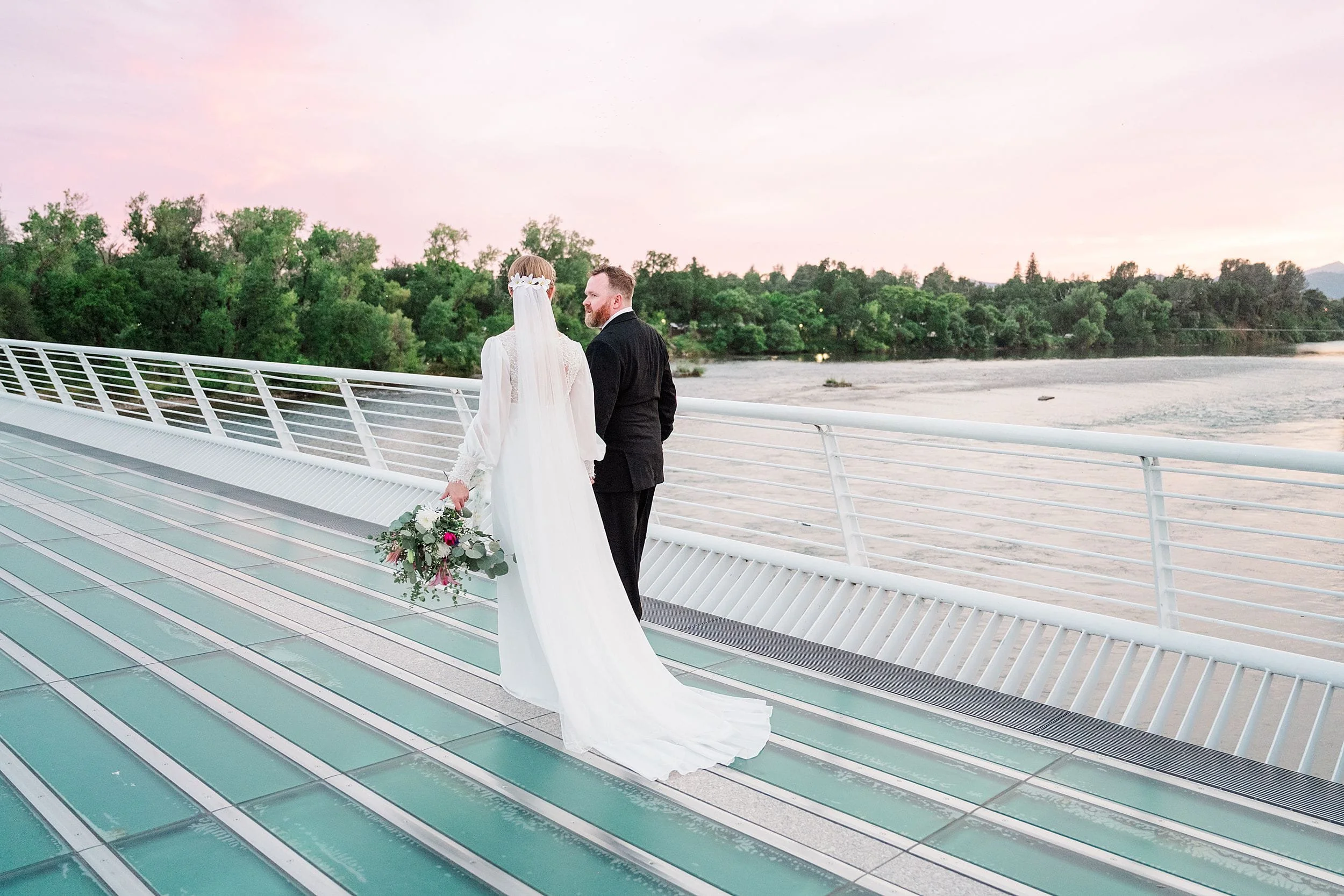 A bride in a long white dress and groom in a black suit stand on a bridge at sunset, holding hands and gazing at the river below, with trees in the background.