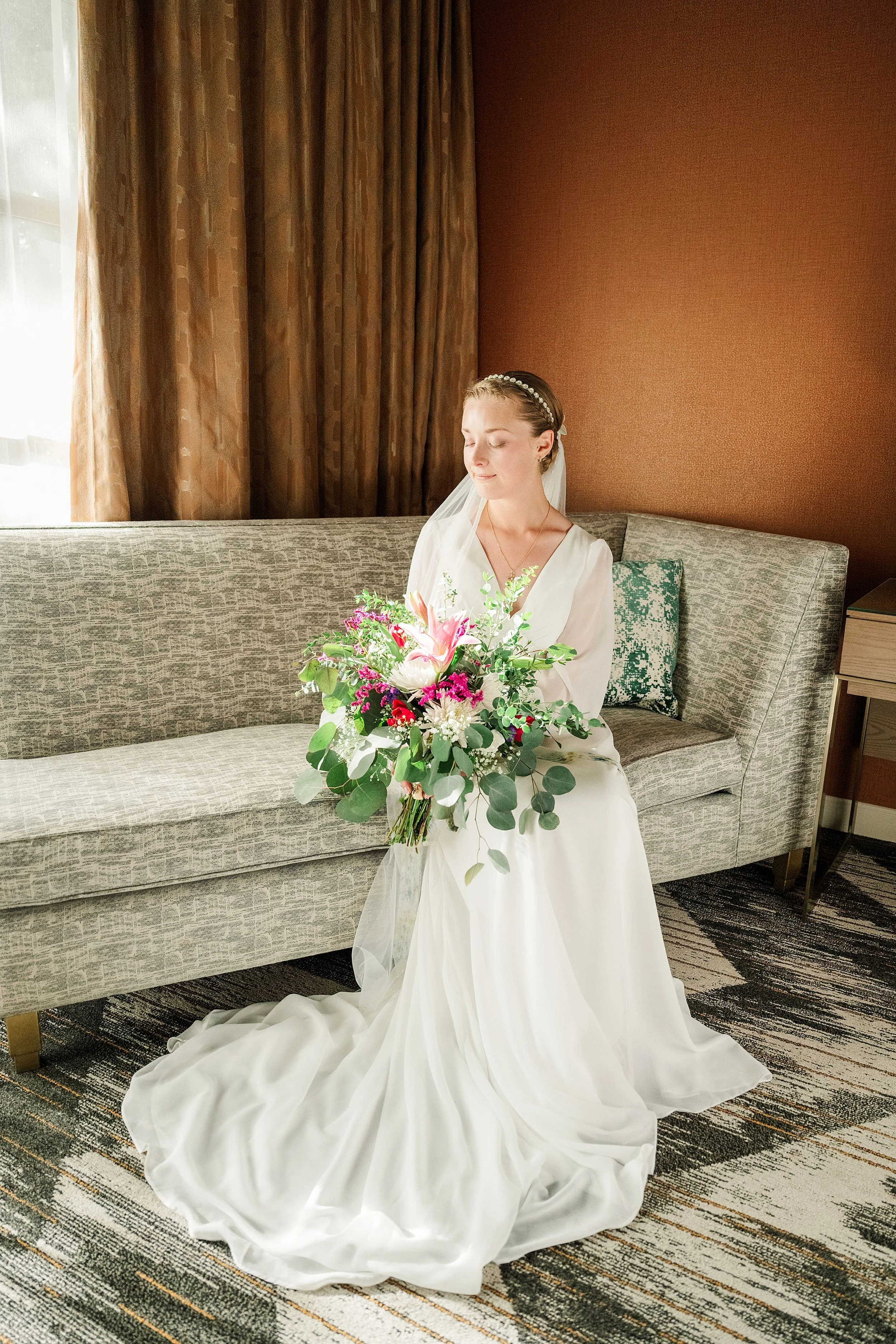 Bride in a white gown sits on a gray sofa, holding a vibrant bouquet of pink and green flowers. The room has warm tones and a sunlight window.