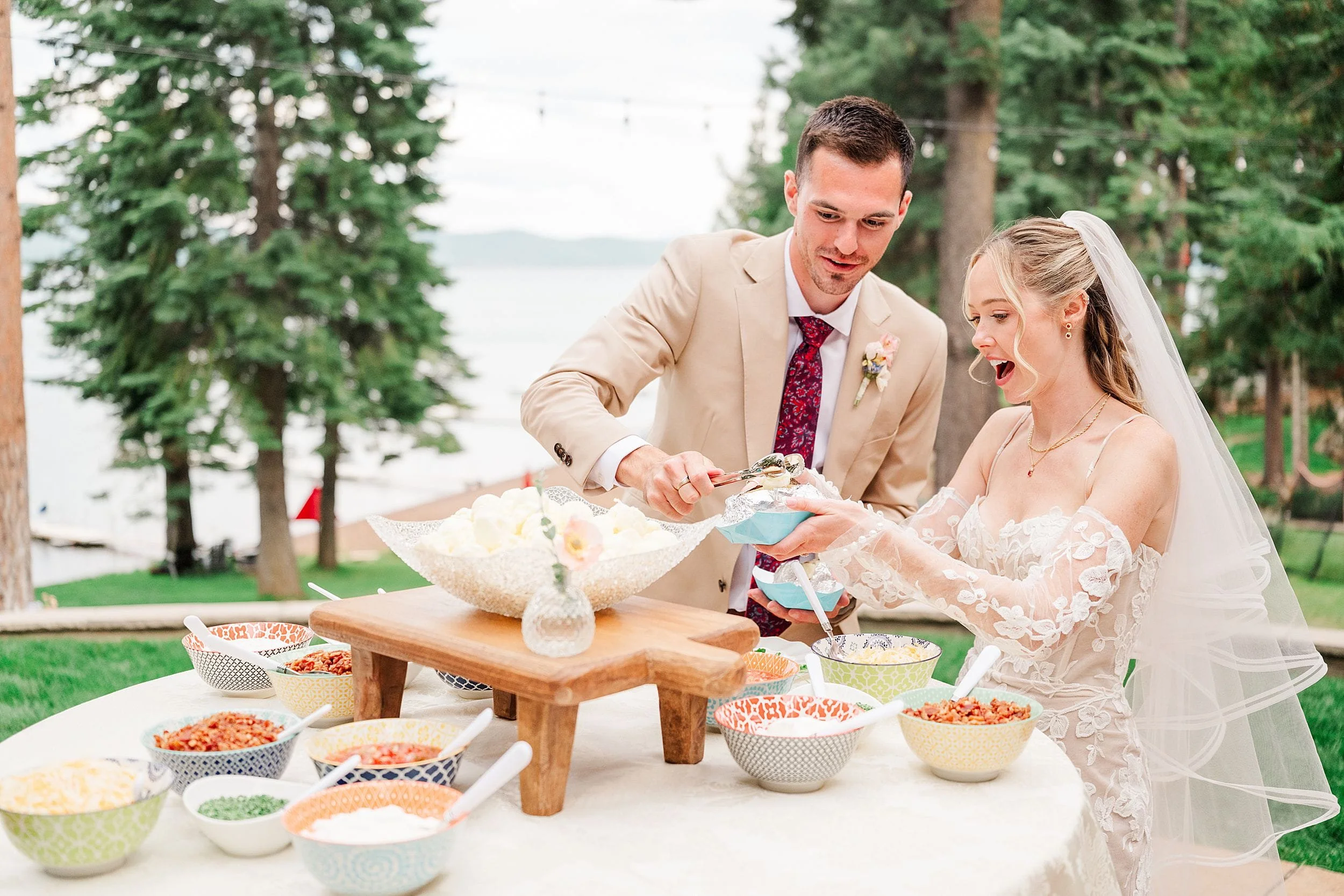Newlywed bride and groom get their food from potato bar at an outdoor wedding reception by a lake, surrounded by pine trees, conveying joy and celebration.
