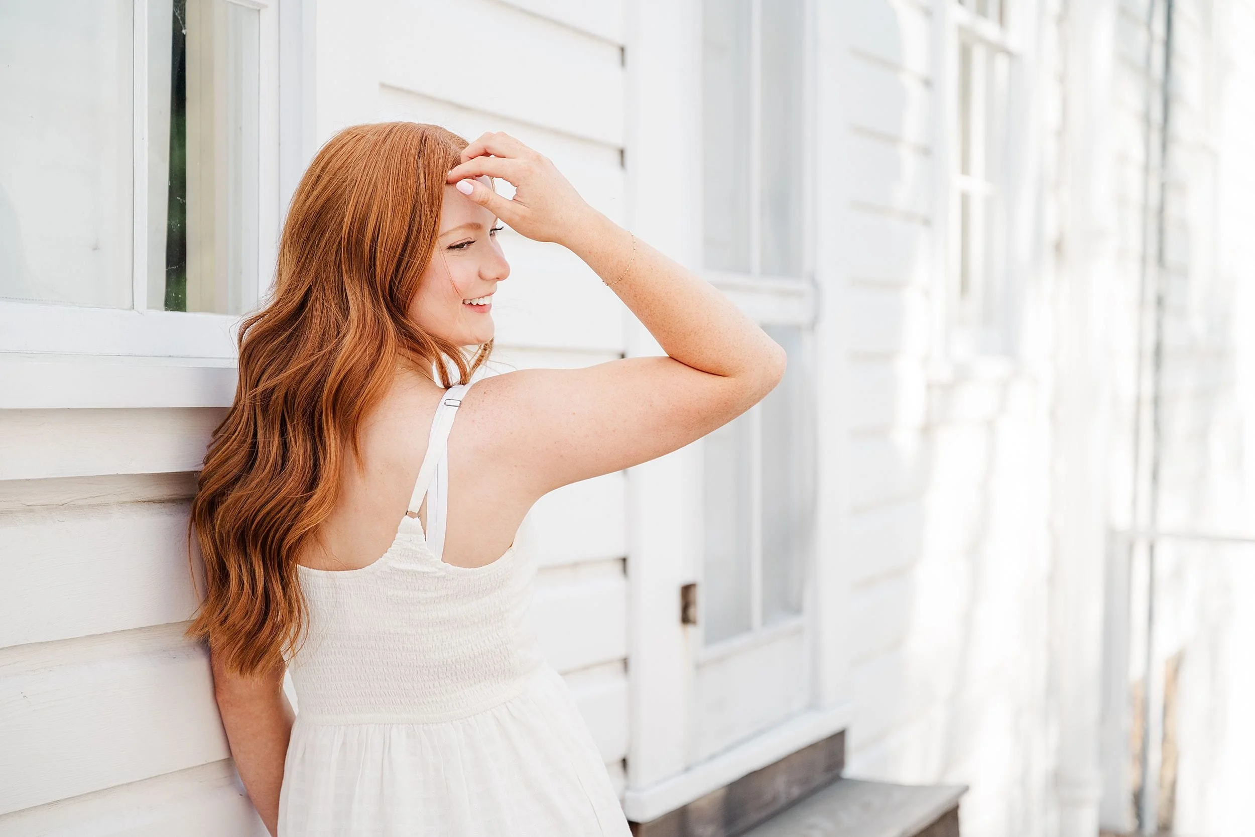 A woman with long red hair leans against a white wooden wall, smiling softly. She wears a white dress, with sunlight creating a warm glow, conveying a serene mood.