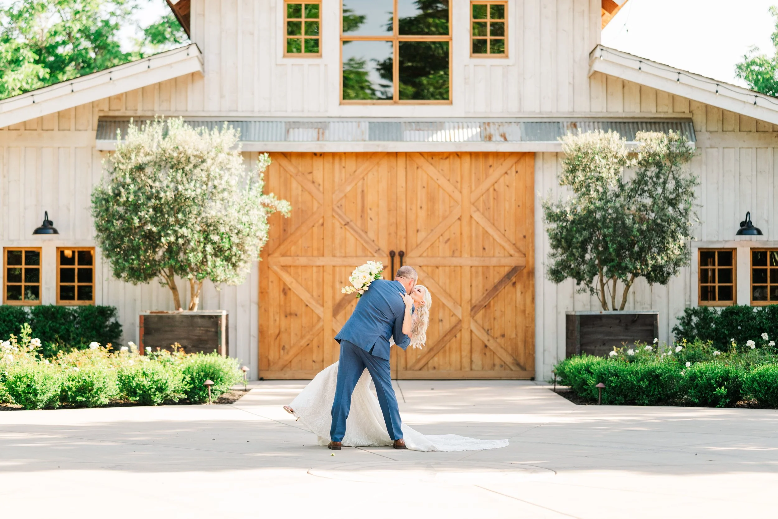 A newlywed couple dancing outside in front of a rustic barn, at The Barn at Pheasant Trail, with large wooden doors, surrounded by greenery and trees, during daytime.