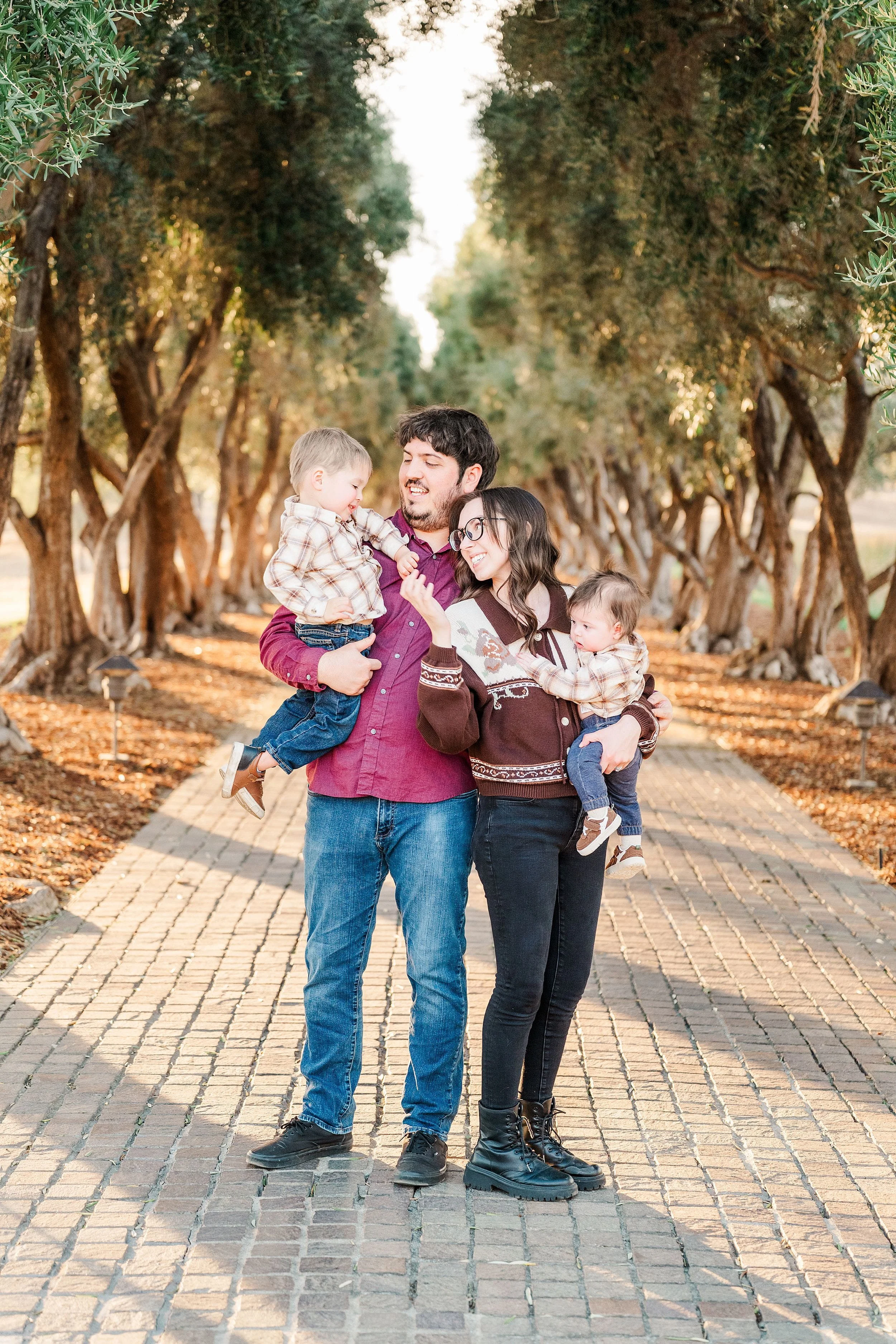 A smiling family of four—parents holding two young children—stands on a sunlit cobblestone path beneath a canopy of trees, sharing a warm moment.