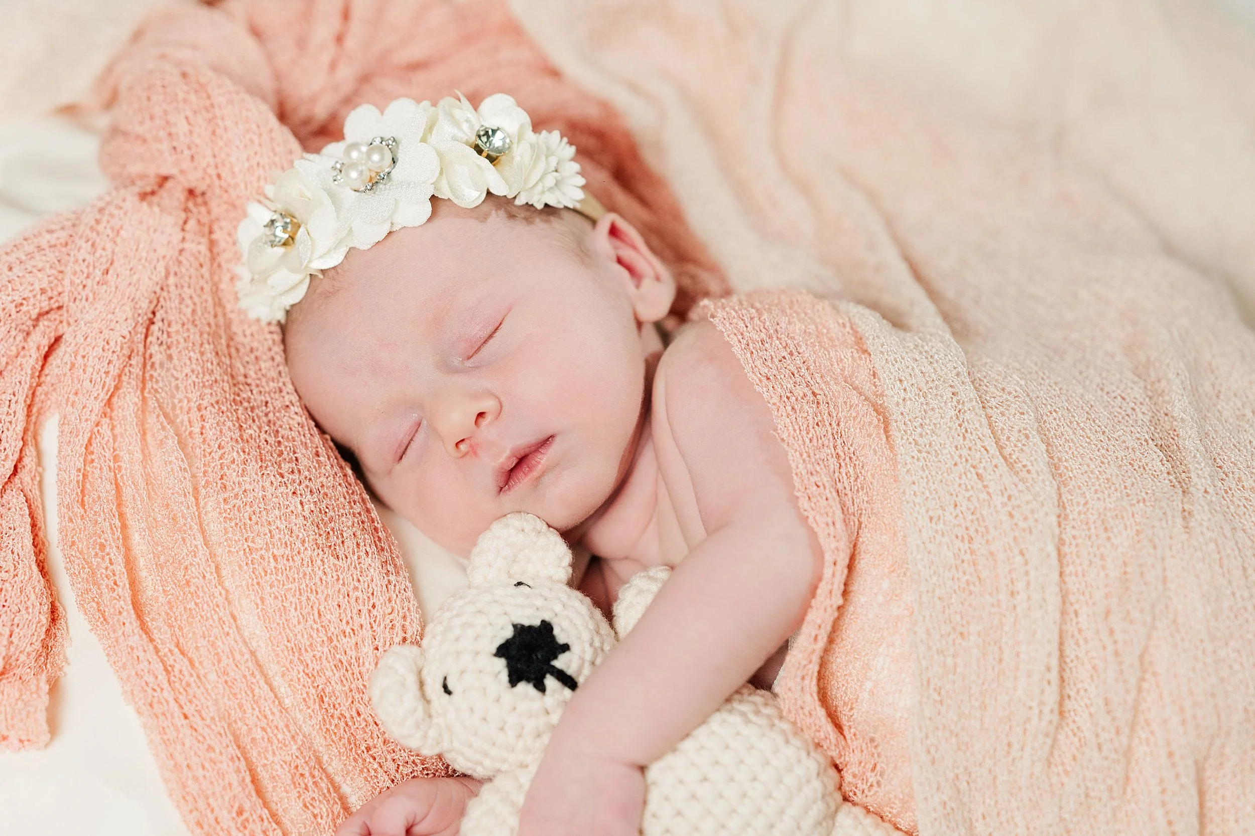 Newborn baby sleeps peacefully on peach fabric, wearing a floral headband, and cuddling a knitted white teddy bear. The scene is serene and tender.