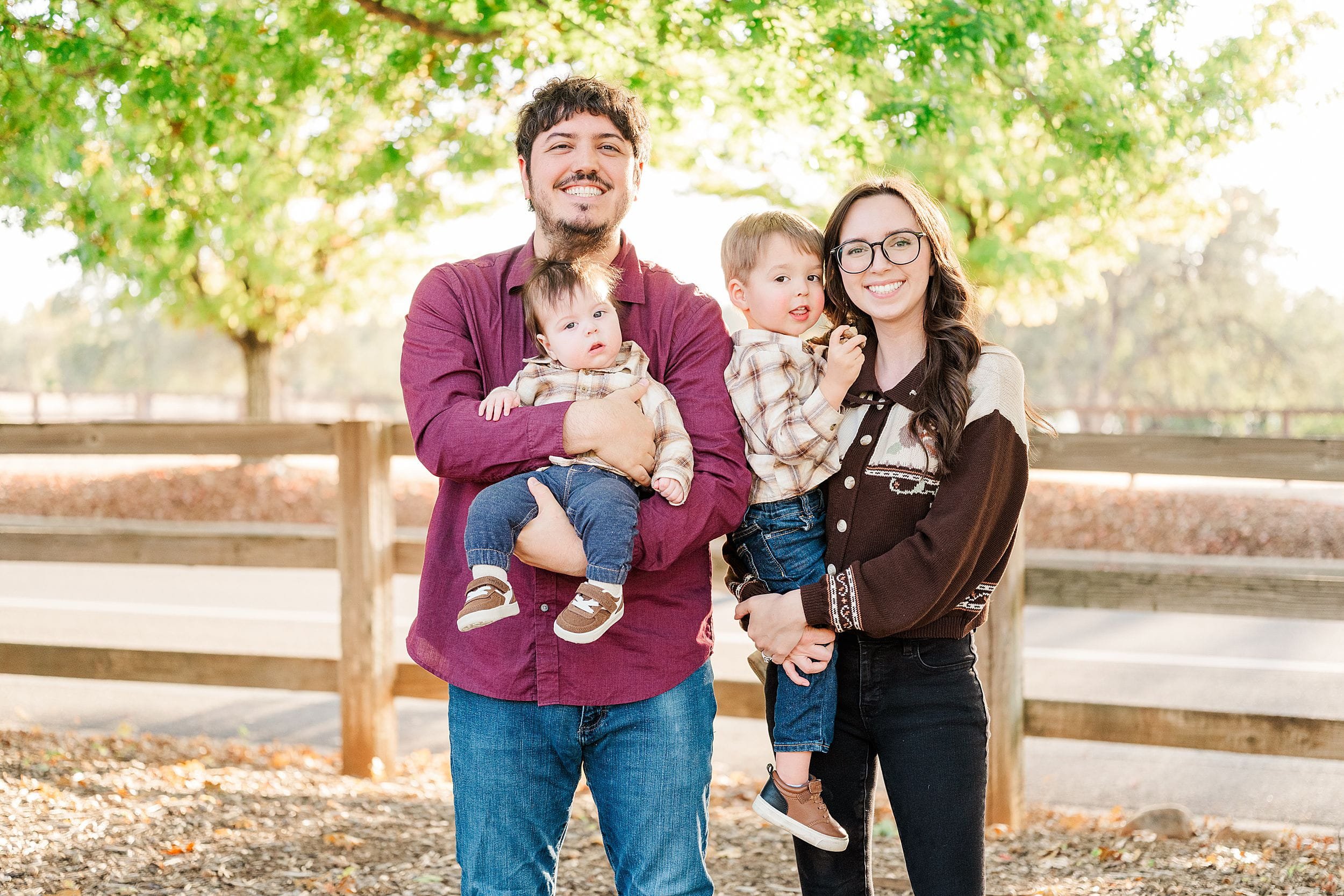 A smiling family of four poses beneath leafy trees by a wooden fence, parents holding their young children in a bright, warm outdoor portrait.