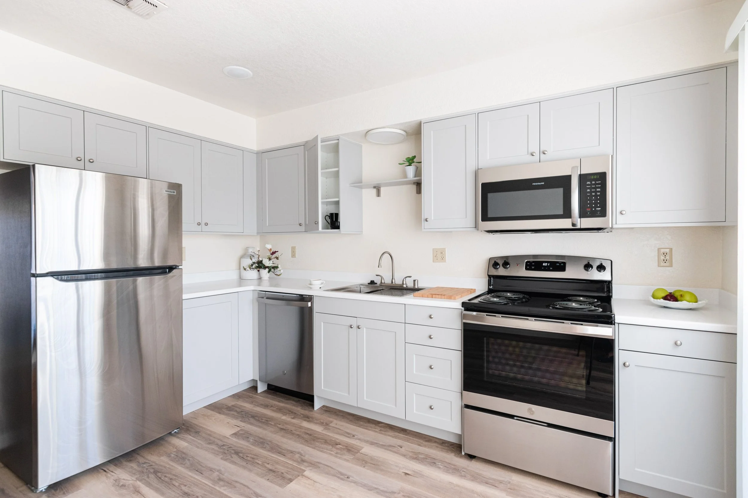 Bright kitchen with white cabinets, stainless steel refrigerator, microwave, stove, dishwasher, and wooden flooring, decorated with a bowl of apples and flowers.
